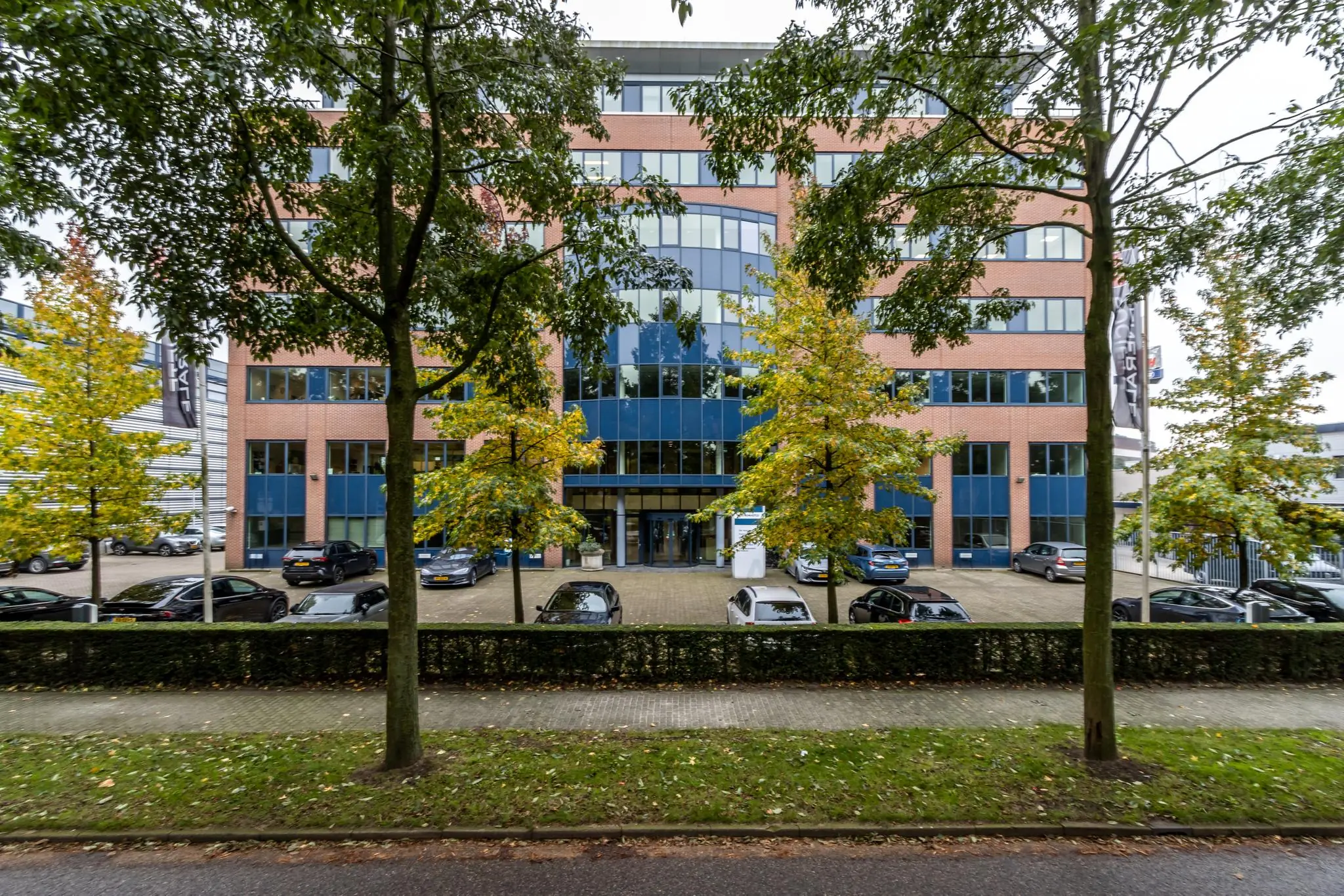 Vooraanzicht van een modern kantoorgebouw met geparkeerde auto's en bomen langs de straat.