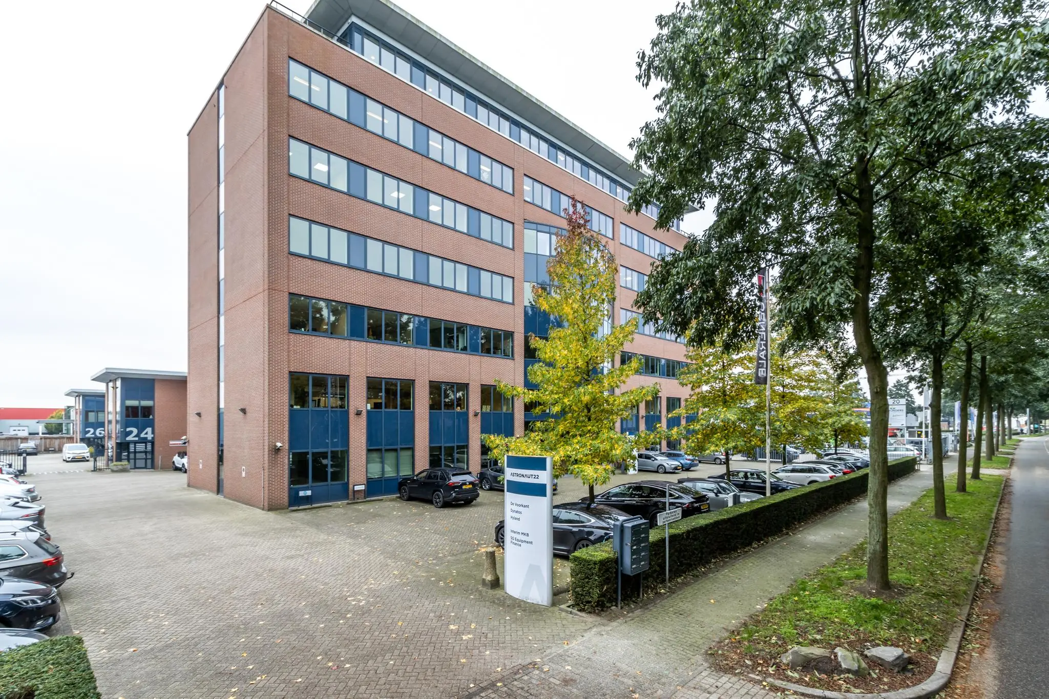 Kantoorgebouw met de naam "Astronaut 24-26" en parkeergelegenheid aan een boomrijke straat.