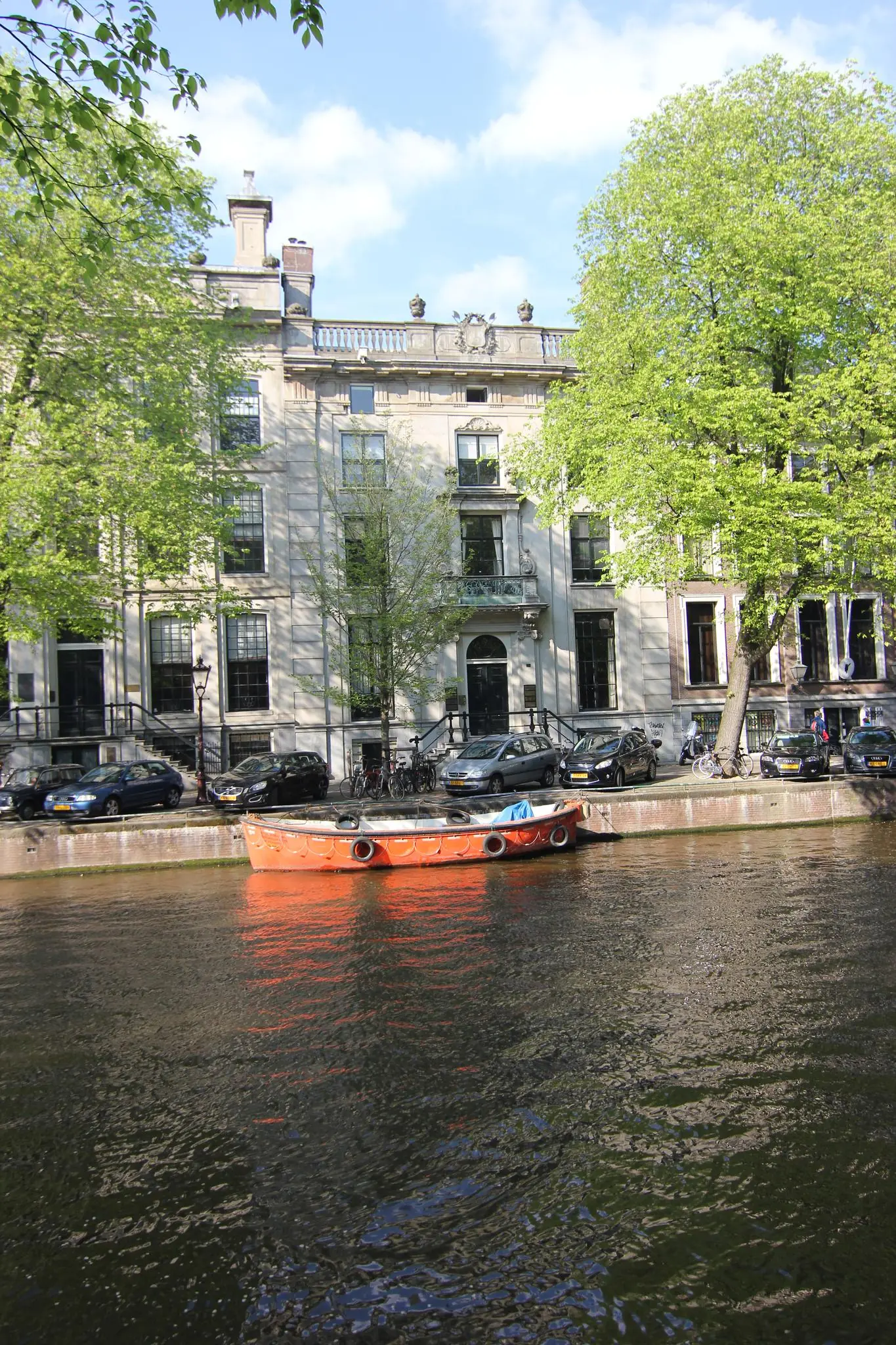 Een oranje boot ligt aangemeerd aan de Herengracht in Amsterdam, met statige grachtenpanden en bomen op de achtergrond.