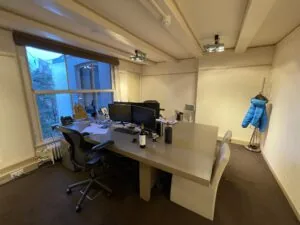 Modern kantoor aan de Herengracht met een groot bureau, twee bureaustoelen, computerschermen en een blauwe jas aan de kapstok.