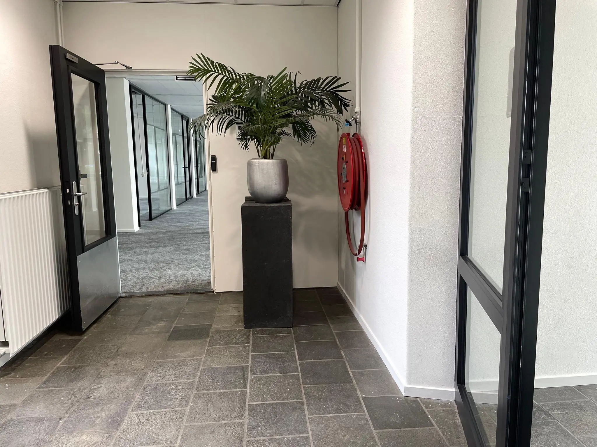 Ingang van een modern kantoorpand aan de Kernkade met een plant op een sokkel en een brandslanghaspel aan de muur.
