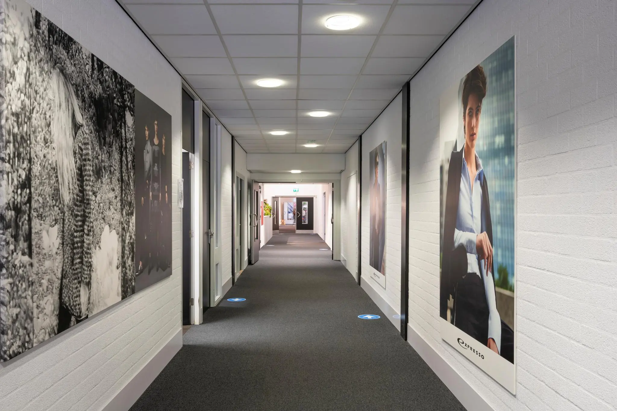 Een verlicht kantoorgebouwgang met grote modeposters aan de muren, waaronder een Expresso-reclame.