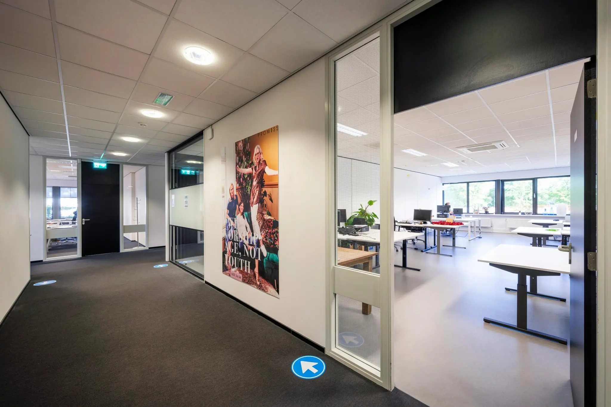 Moderne kantoorruimte met open werkplekken, grote ramen en een poster aan de muur in de gang met de tekst "WORSHIP AGE, NOT YOUTH".
