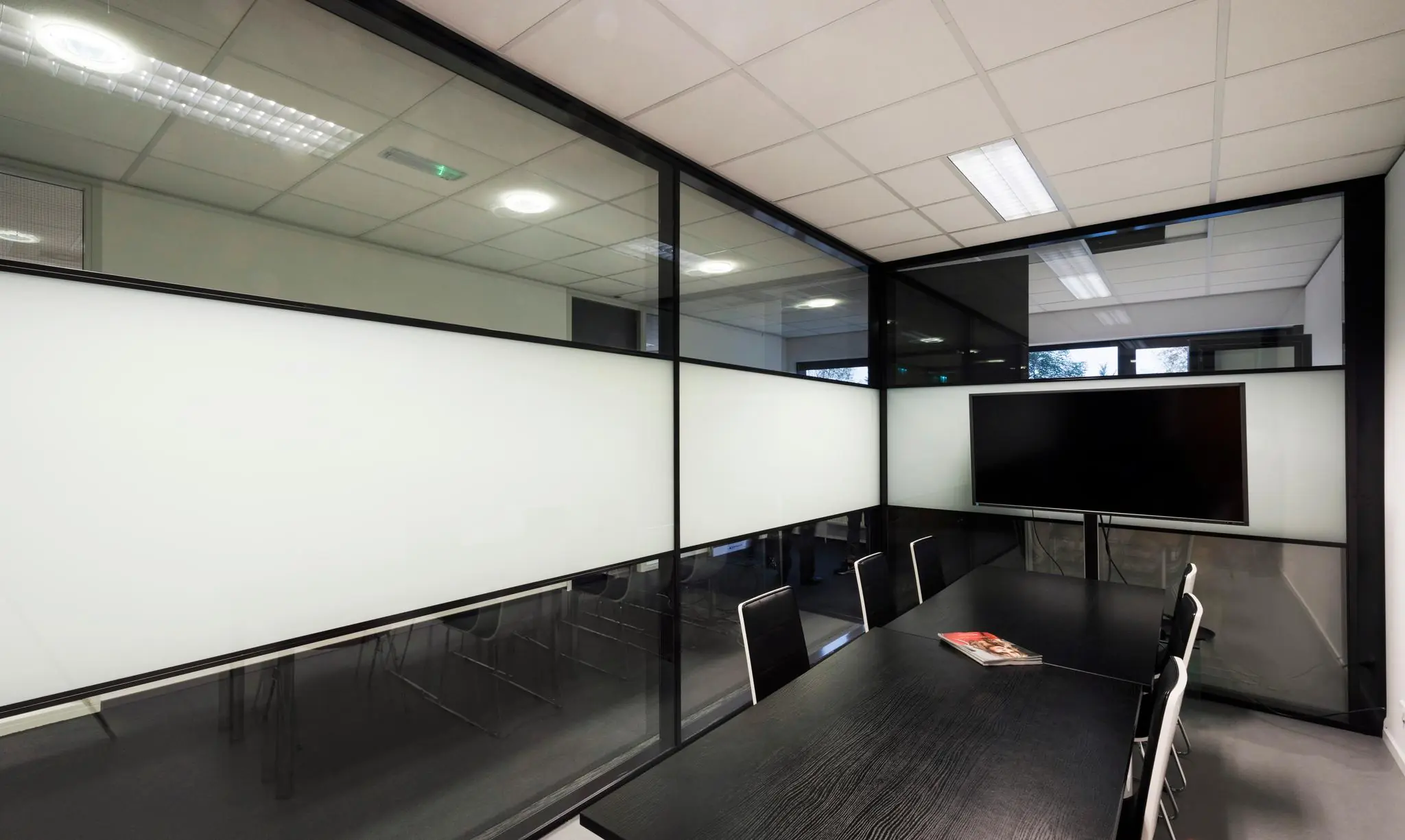 Vergaderruimte met zwarte tafel, moderne stoelen, glazen wanden en een groot scherm aan de muur.