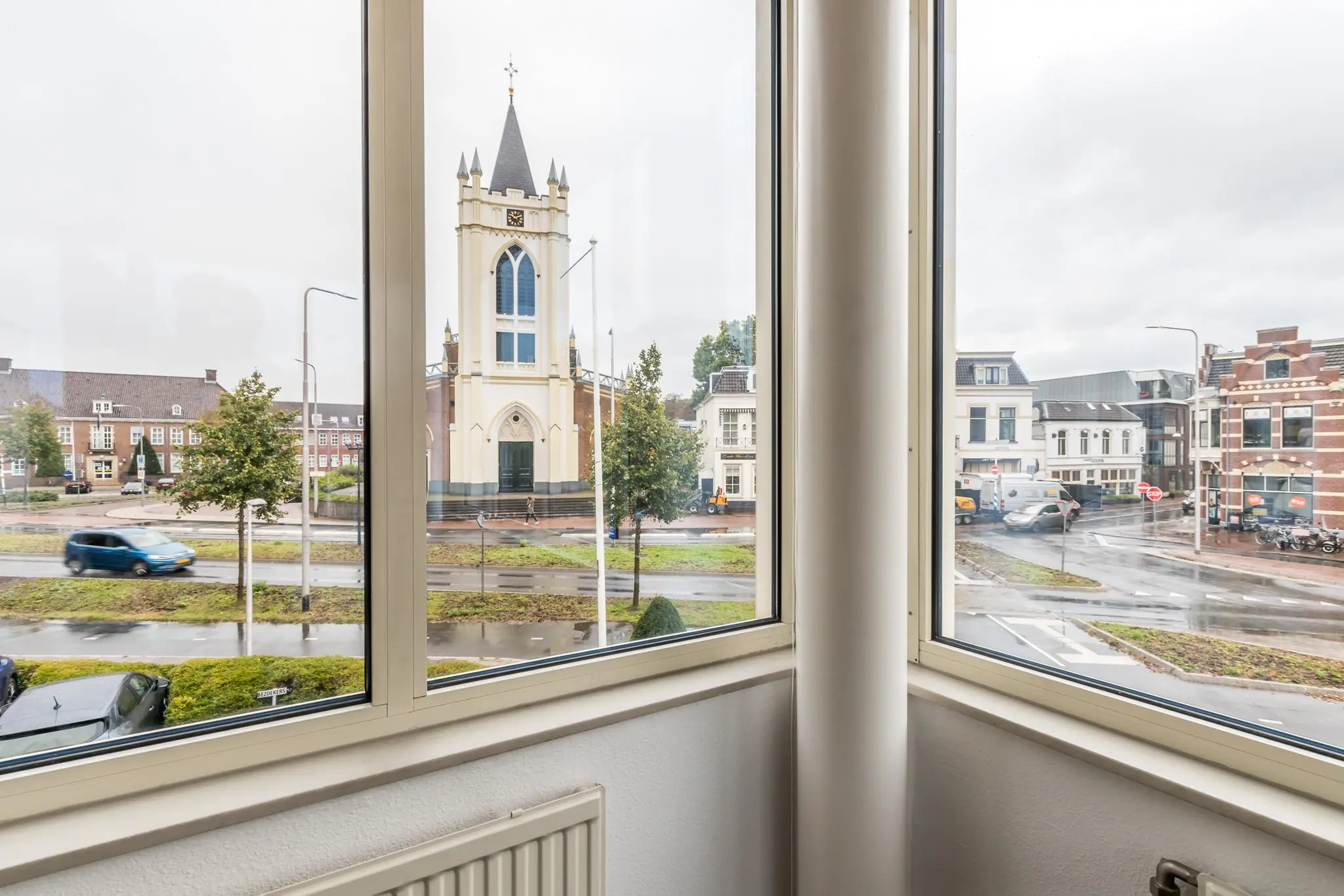 Uitzicht vanuit een raam op de Utrechtseweg met een kerk en omliggende bebouwing op een regenachtige dag.