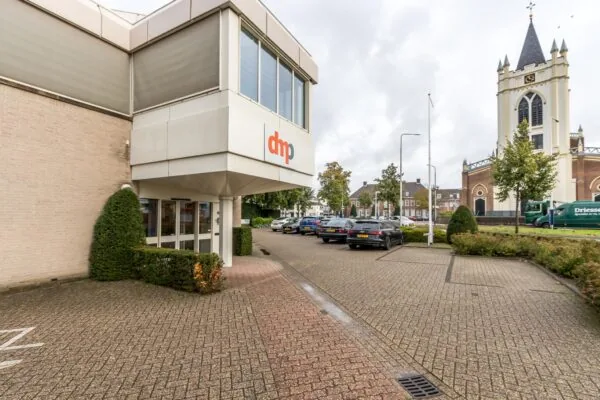 Ingang van het kantoorpand van DMP aan de Utrechtseweg met op de achtergrond een kerk en geparkeerde auto's.