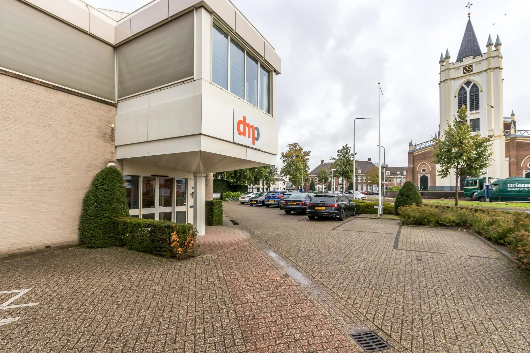 Ingang van het kantoorpand van DMP aan de Utrechtseweg met op de achtergrond een kerk en geparkeerde auto's.