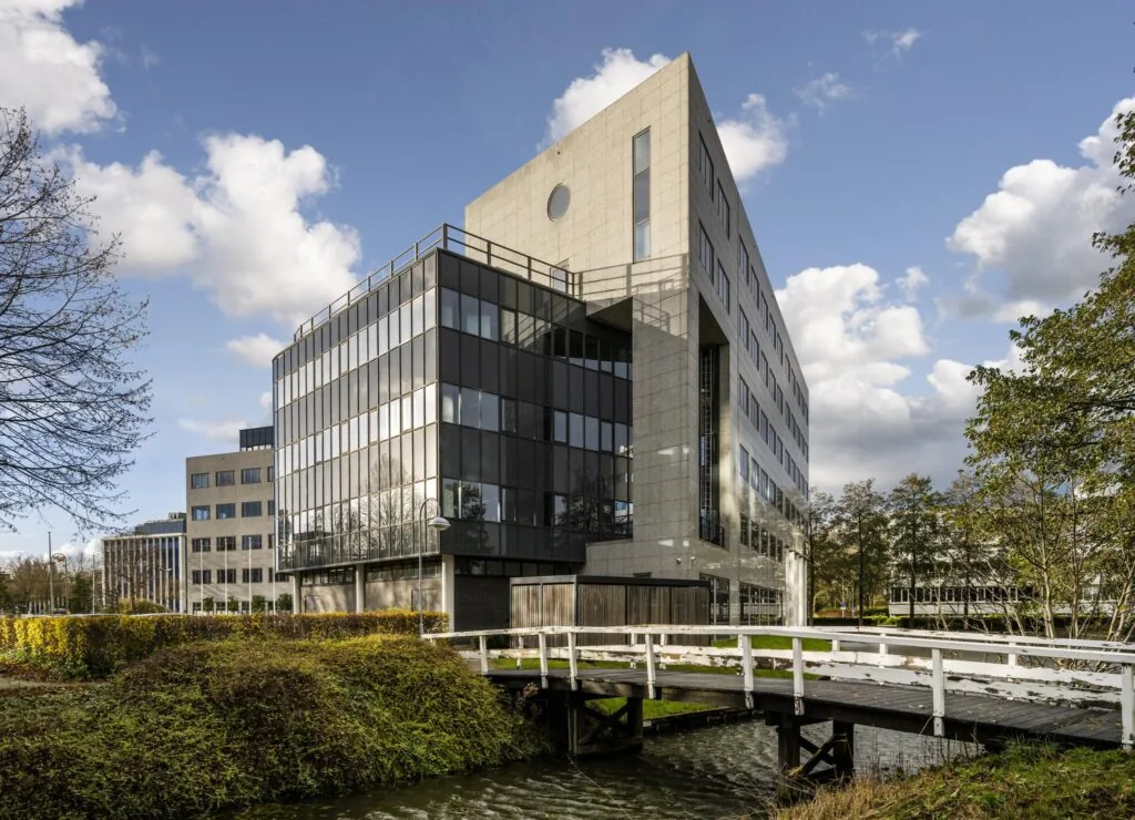 Modern kantoorgebouw aan de Neptunusstraat met glazen gevel en wit bruggetje over het water op de voorgrond.