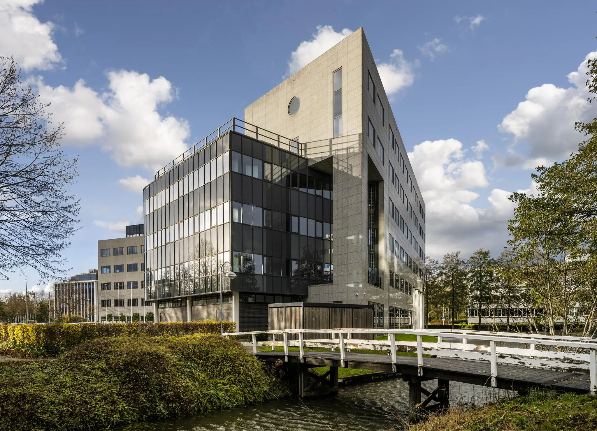 Modern kantoorgebouw aan de Neptunusstraat met glazen gevel en wit bruggetje over het water op de voorgrond.