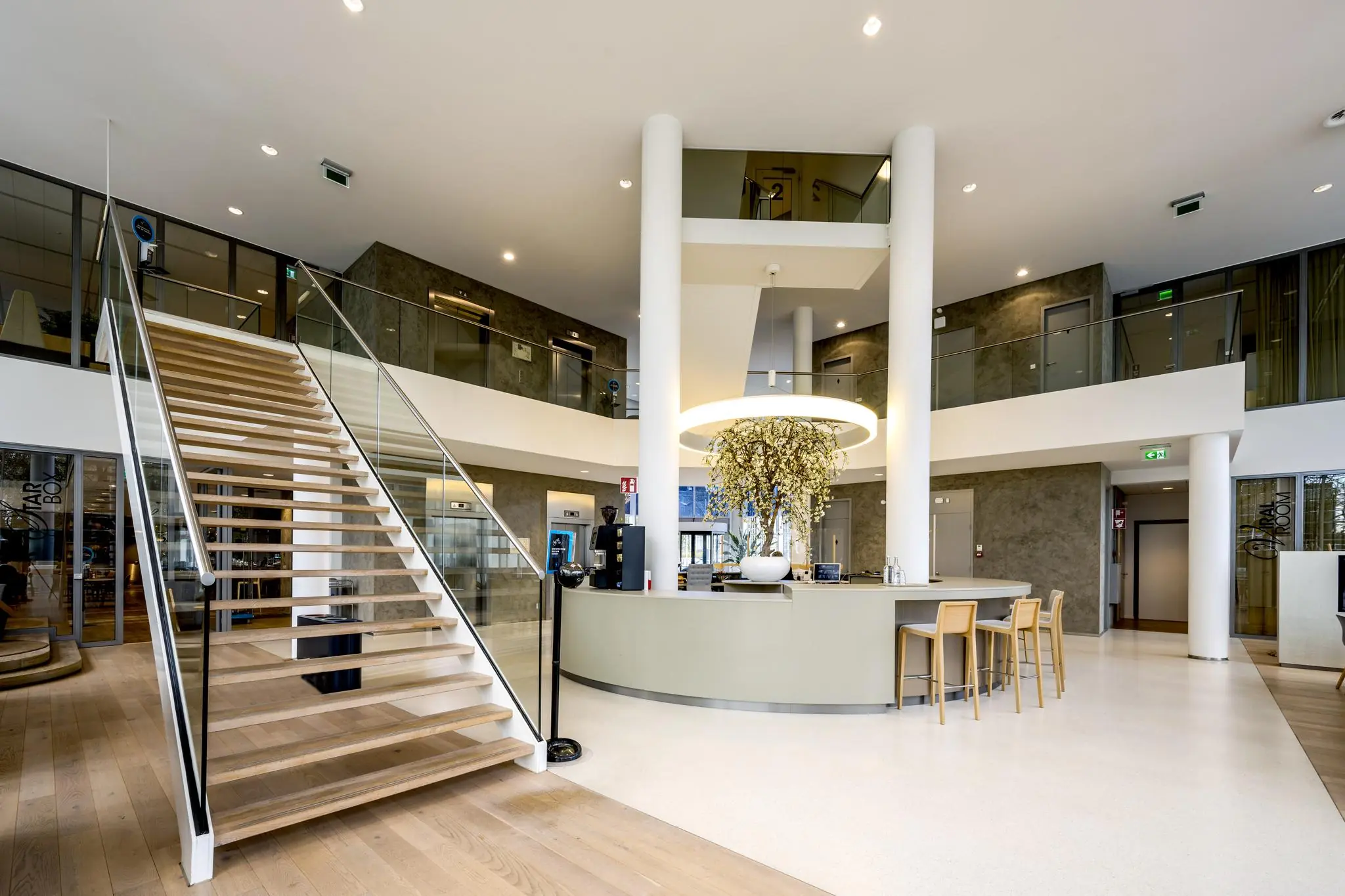 Moderne, ruimtelijke ontvangsthal aan de Neptunusstraat met een houten trap, centraal informatiepunt en stijlvolle inrichting.