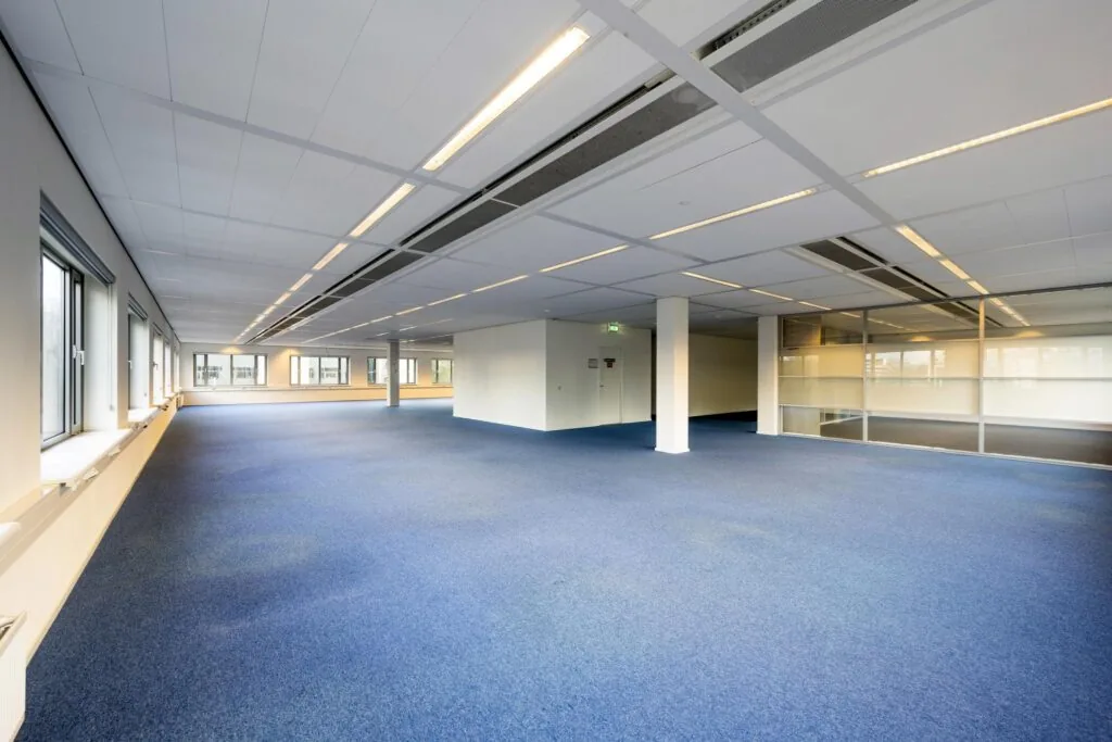 Lichte, lege kantoorruimte aan de Neptunusstraat met blauwe vloerbedekking en grote ramen.