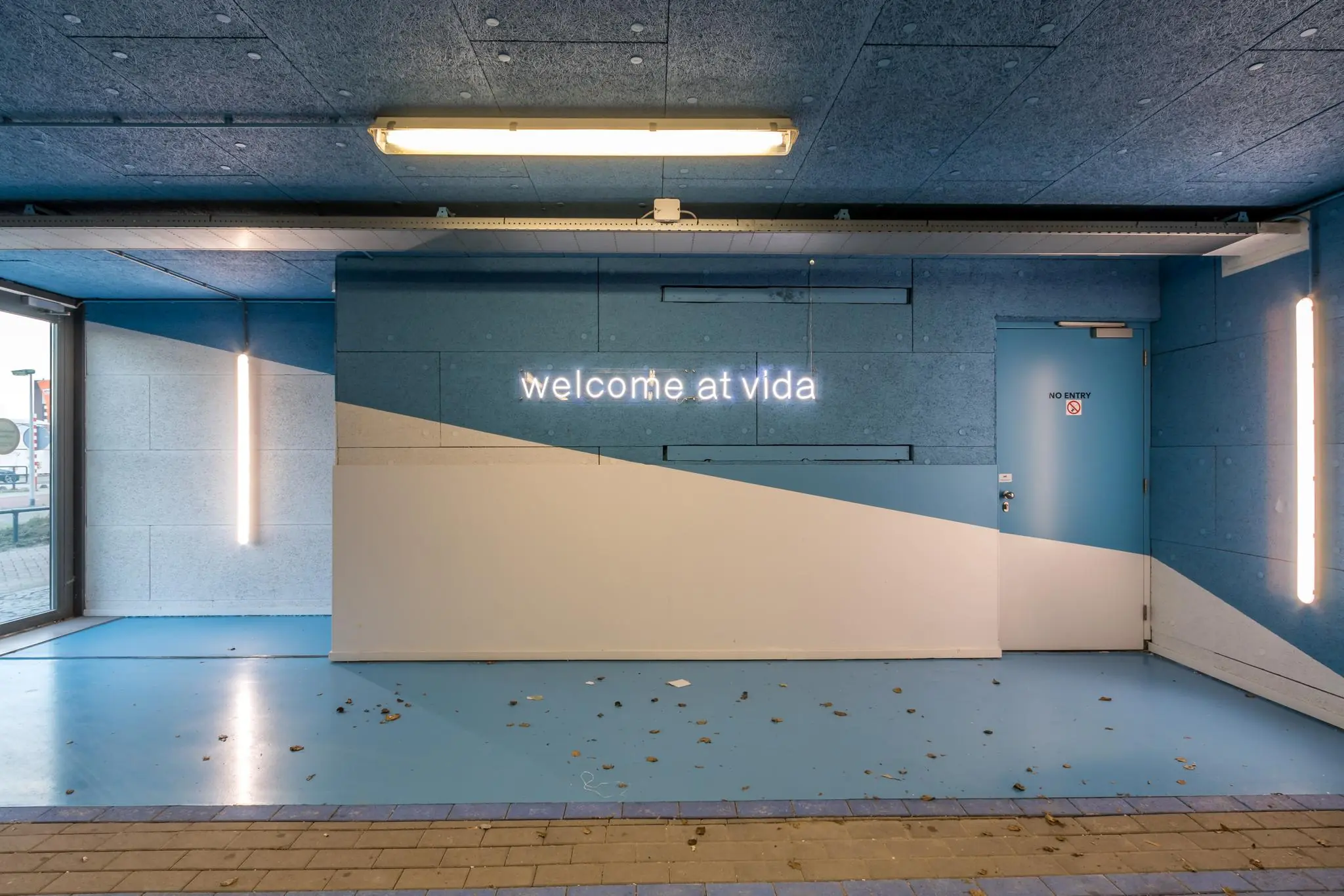 Ingangshal met blauwe en witte accenten, neontekst "welcome at vida" en een deur met "no entry" aanduiding.