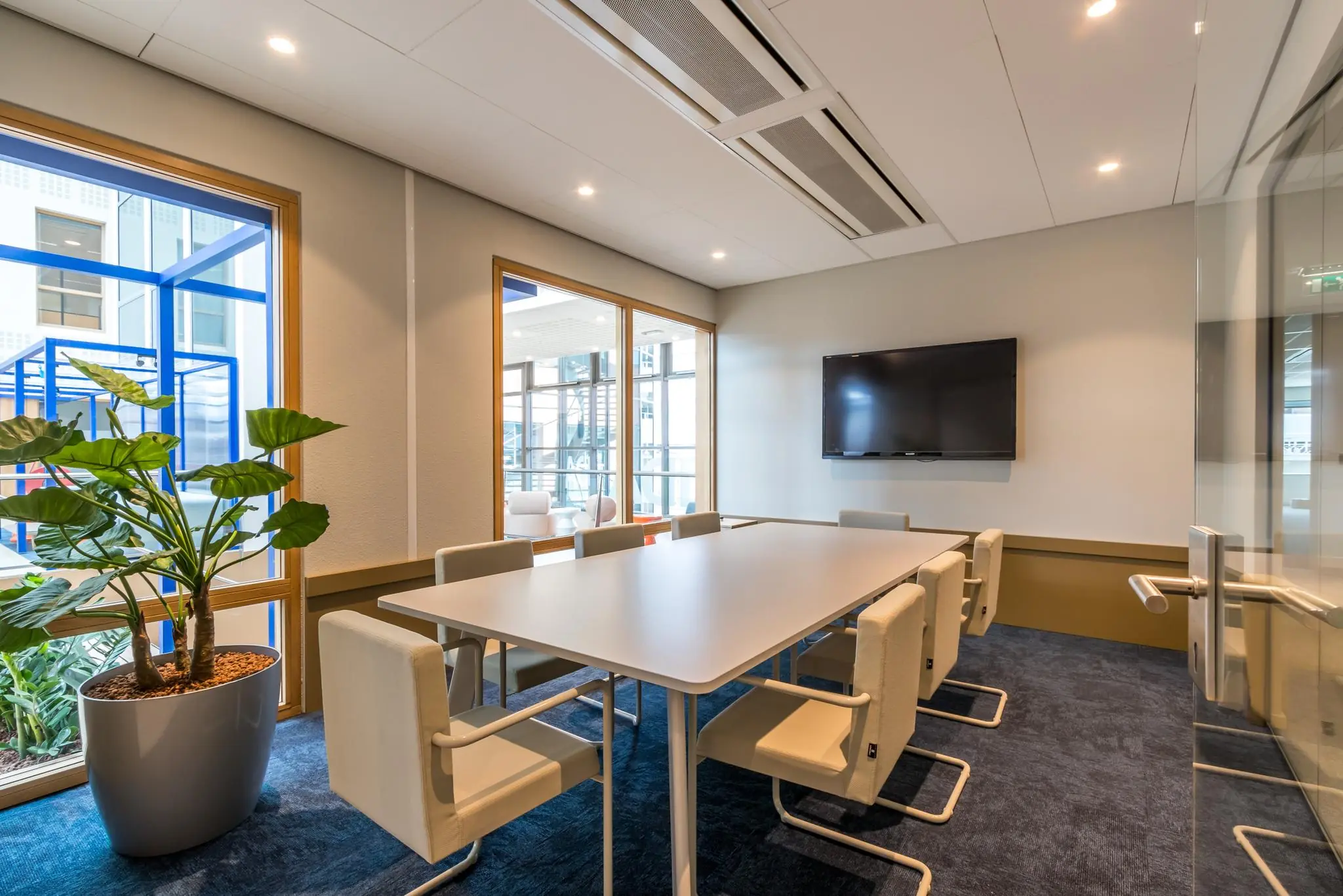Vergaderruimte met moderne inrichting, een lange tafel, acht stoelen, een flatscreen aan de muur en grote ramen met uitzicht op een overdekte binnenruimte.