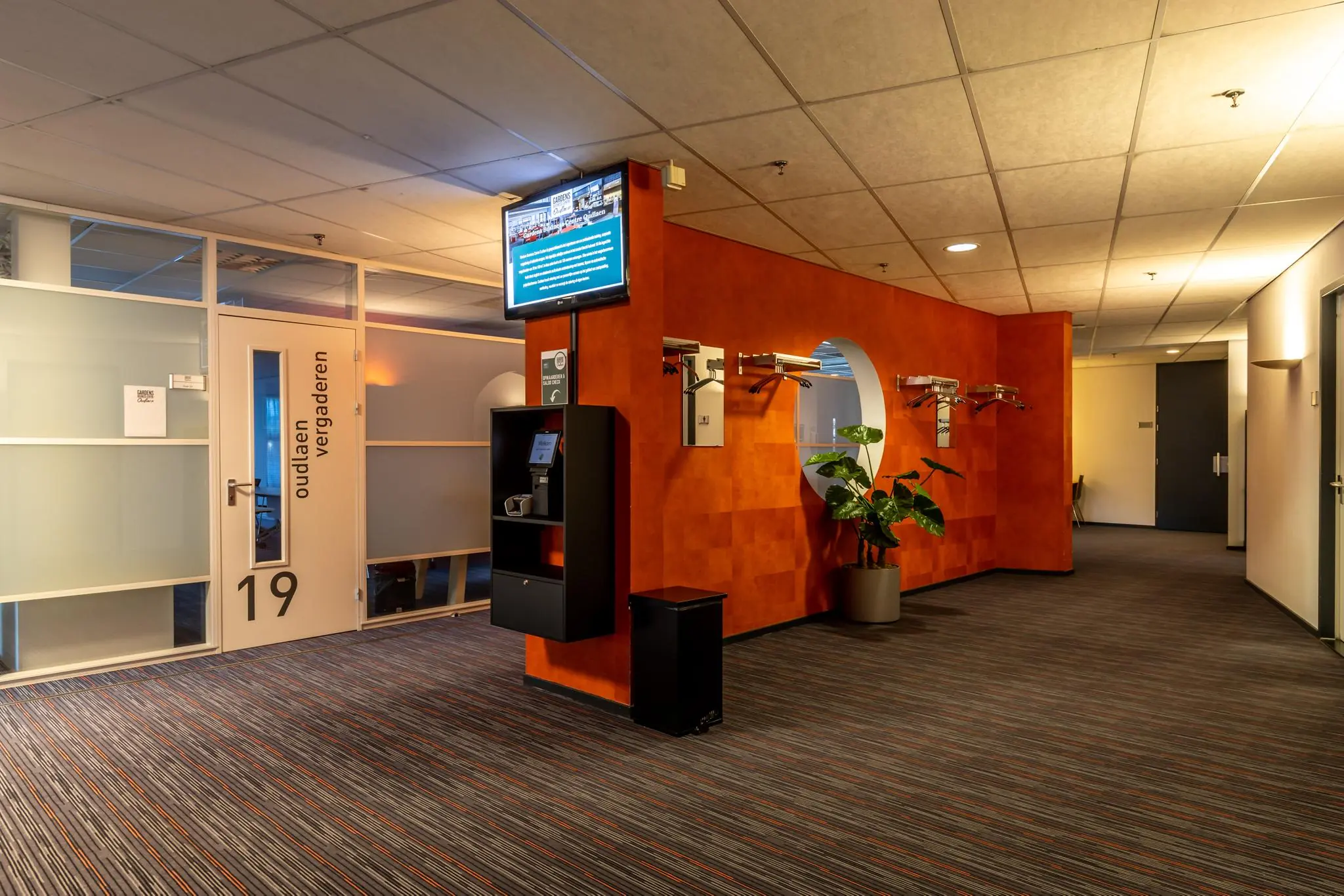 Hal met vergaderruimtes in het gebouw Oudlaan, met een oranje wand, kapstok, digitale informatiezuil en deur naar kamer 19.