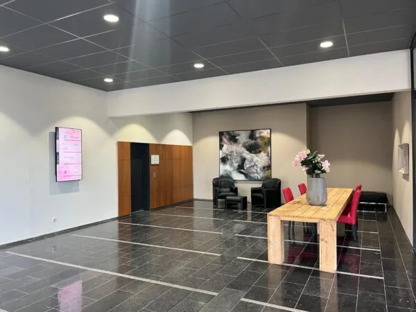 Moderne wachtruimte aan de Arnhemseweg met houten tafel, rode stoelen, kunst aan de muur en een digitale informatiezuil.