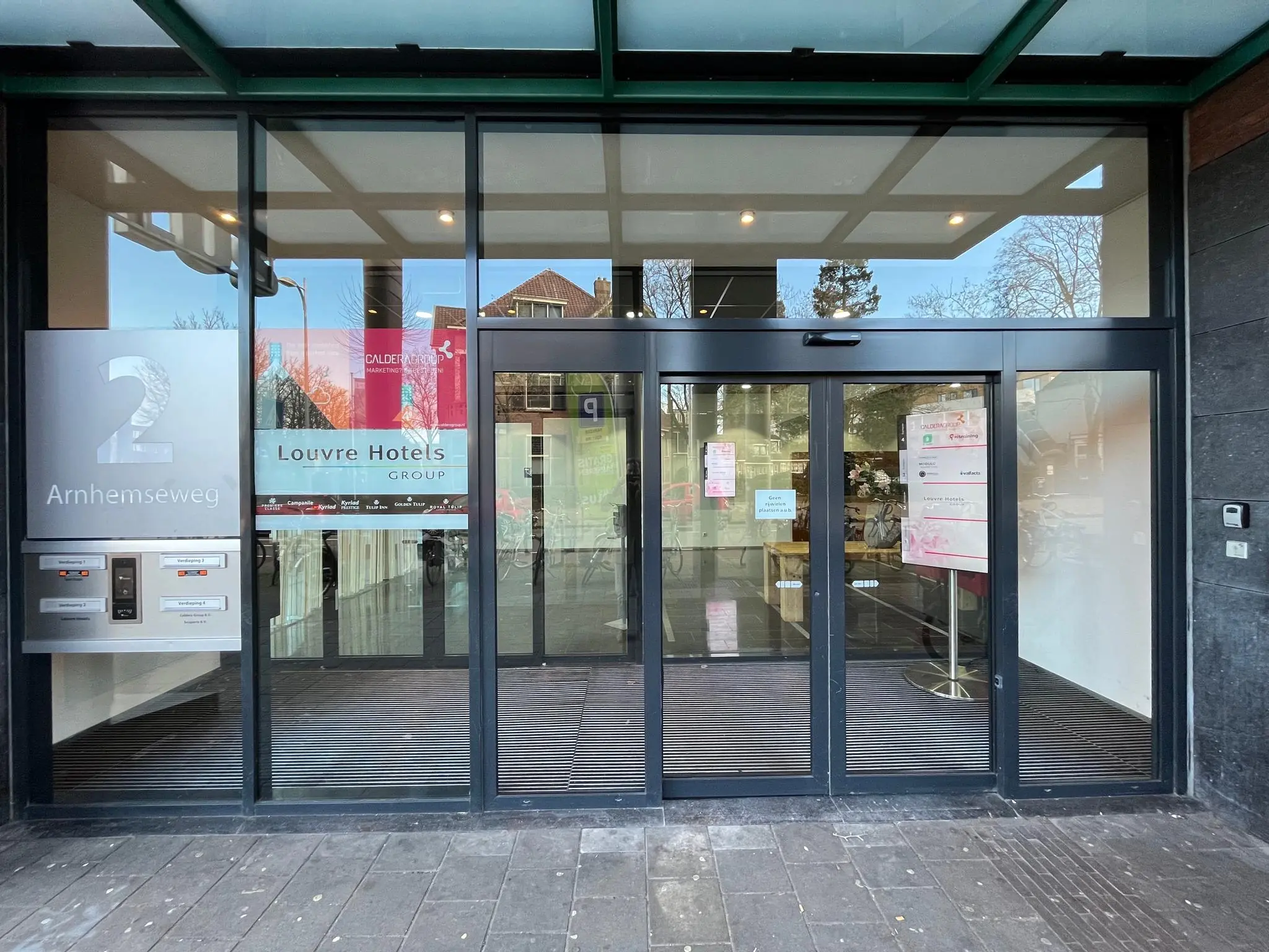 Ingang van het gebouw aan de Arnhemseweg 2 met automatische schuifdeuren en signage van de Louvre Hotels Group.