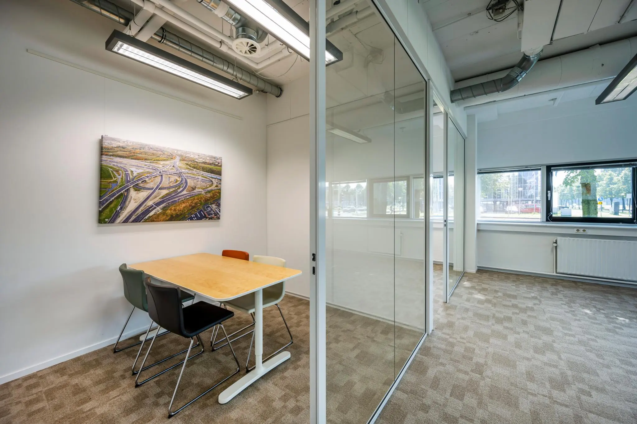 Vergaderruimte met een houten tafel, vier stoelen en een wandfoto van knooppunt Hogehilweg, gescheiden door een glazen wand van een leeg kantoor.