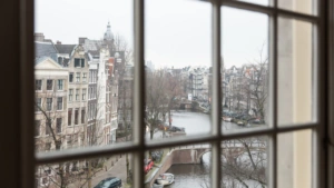 Uitzicht door een raam op de Keizersgracht in Amsterdam met grachtenpanden, brug en water.