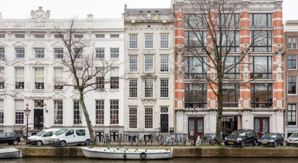 Gevels van historische grachtenpanden aan de Keizersgracht in Amsterdam met geparkeerde auto’s, fietsen en een bootje op de voorgrond.
