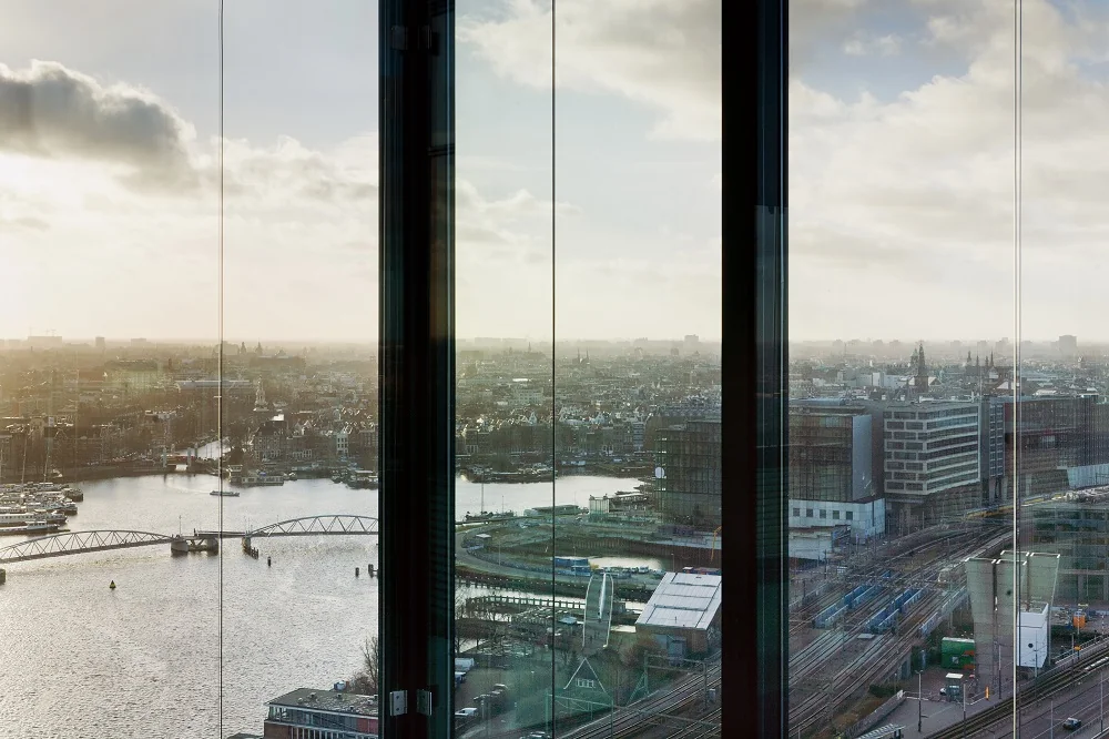 Uitzicht over Amsterdam vanaf de Piet Heinkade, met zicht op het IJ, spoorrails en stedelijke bebouwing bij zonsopkomst.