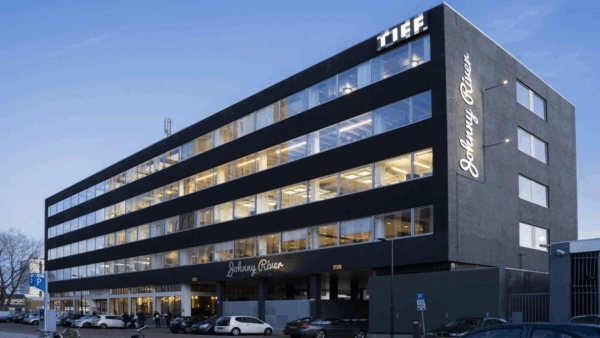 Modern zwart kantoorgebouw aan de Joan Muyskenweg met verlichte gevelletters "Johnny River" en "T.I.E.F." bij schemering.