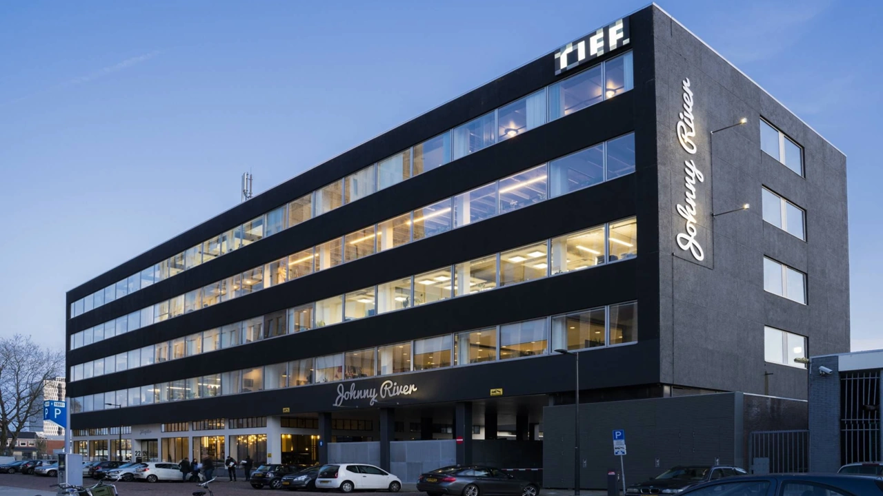 Modern zwart kantoorgebouw aan de Joan Muyskenweg met verlichte gevelletters "Johnny River" en "T.I.E.F." bij schemering.