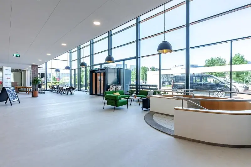 Moderne, lichte ontvangsthal aan de Polarisavenue met grote ramen, groene zitmeubels en een trap naar beneden.