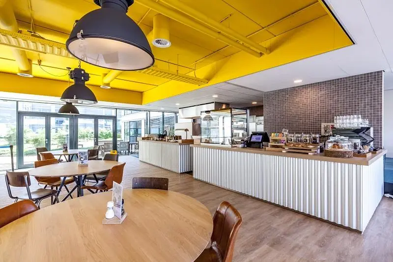 Moderne en lichte kantine aan de Polarisavenue met houten tafels, industriële lampen en een gele plafondconstructie.