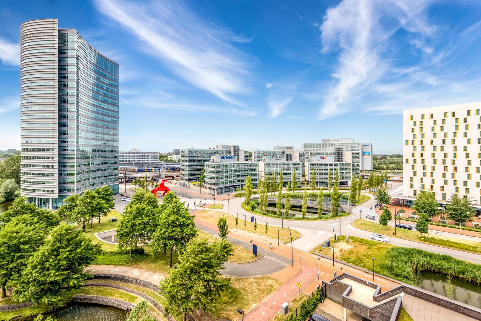 Modern kantorencomplex Scorpius in Amsterdam-Zuidas met omliggende wegen, groen en waterpartijen onder een heldere blauwe lucht.