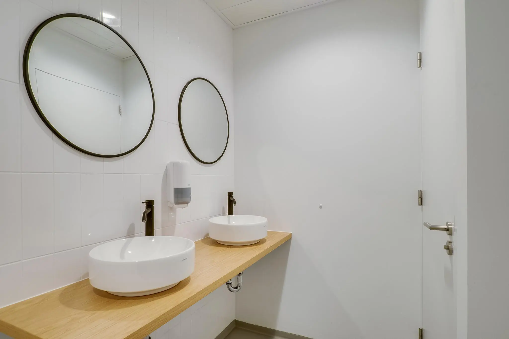 Moderne badkamer met twee ronde spiegels, waskommen op een houten blad en een witte zeepdispenser aan de muur.