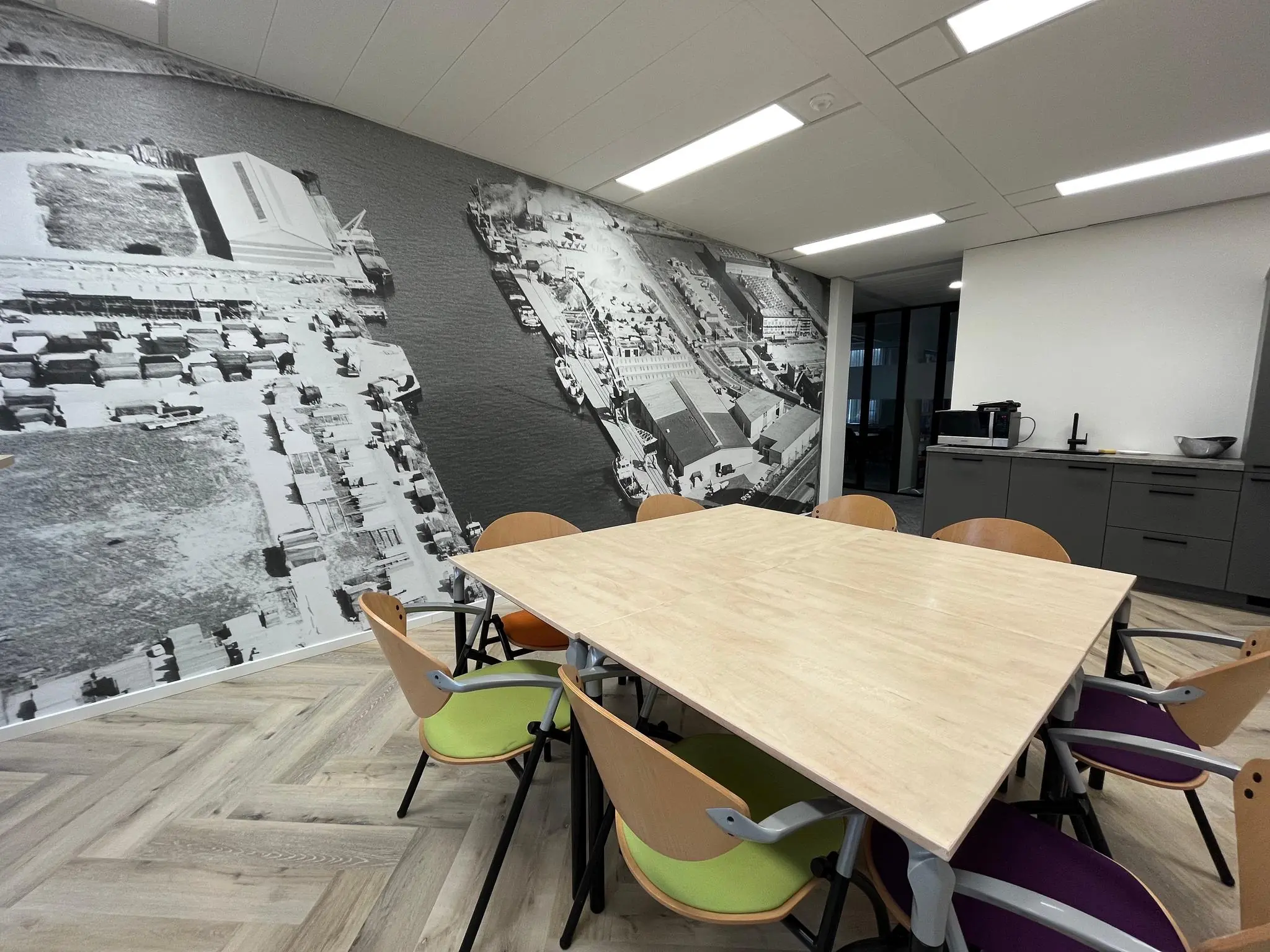 Vergaderruimte met houten tafel, gekleurde stoelen en een muurprint van een oude luchtfoto van de Kernkade.