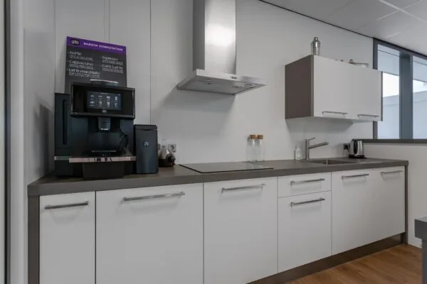 Moderne kantoorkeuken met een koffieautomaat, inductiekookplaat, gootsteen en witte kasten.