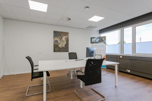 Modern kantoor met een wit bureau, zwarte stoelen, een computerscherm, grote ramen en een schilderij van een zwaan aan de muur.