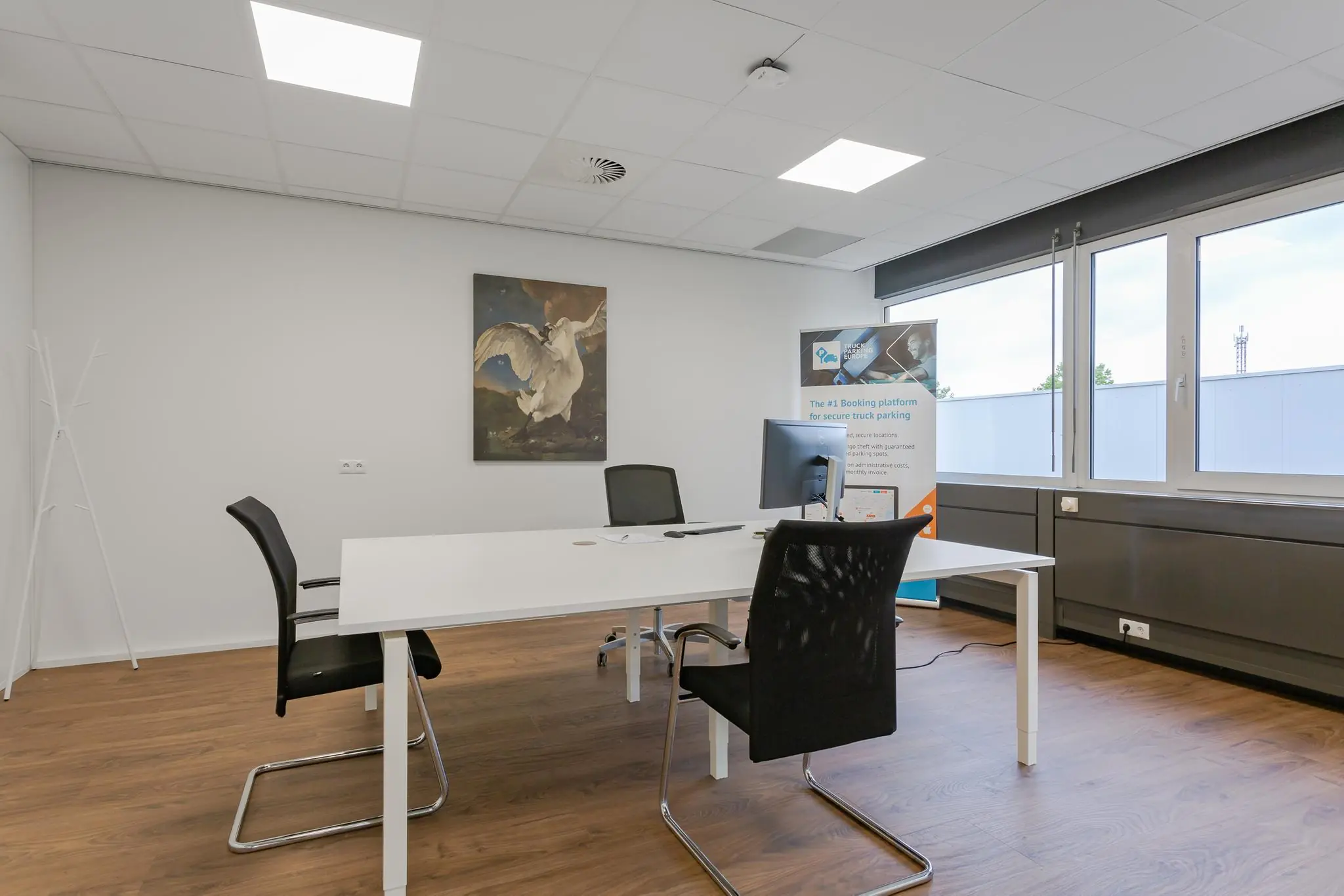 Modern kantoor met een wit bureau, zwarte stoelen, een computerscherm, grote ramen en een schilderij van een zwaan aan de muur.