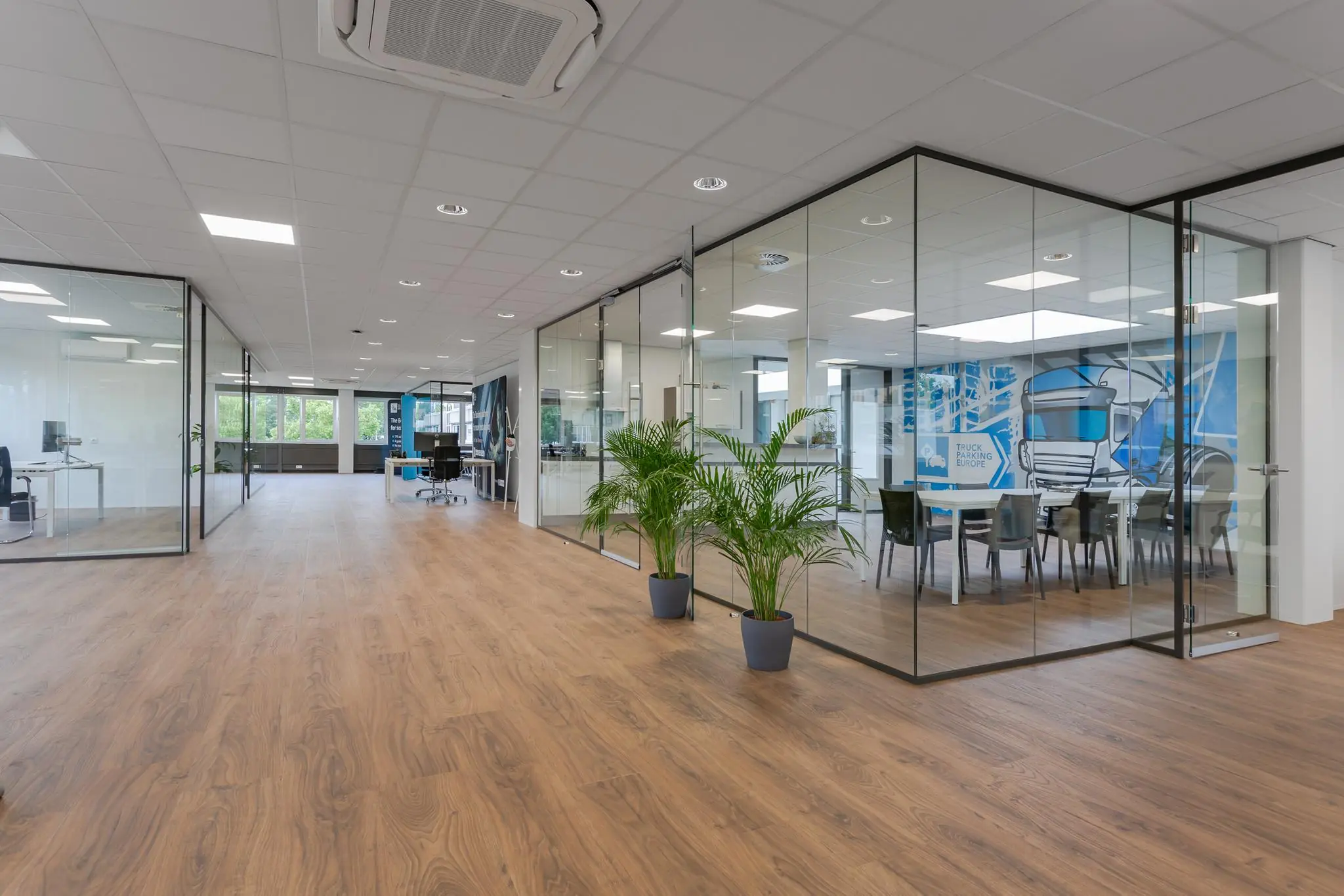 Modern kantoorinterieur met glazen vergaderruimtes, houten vloer en planten langs de gang.