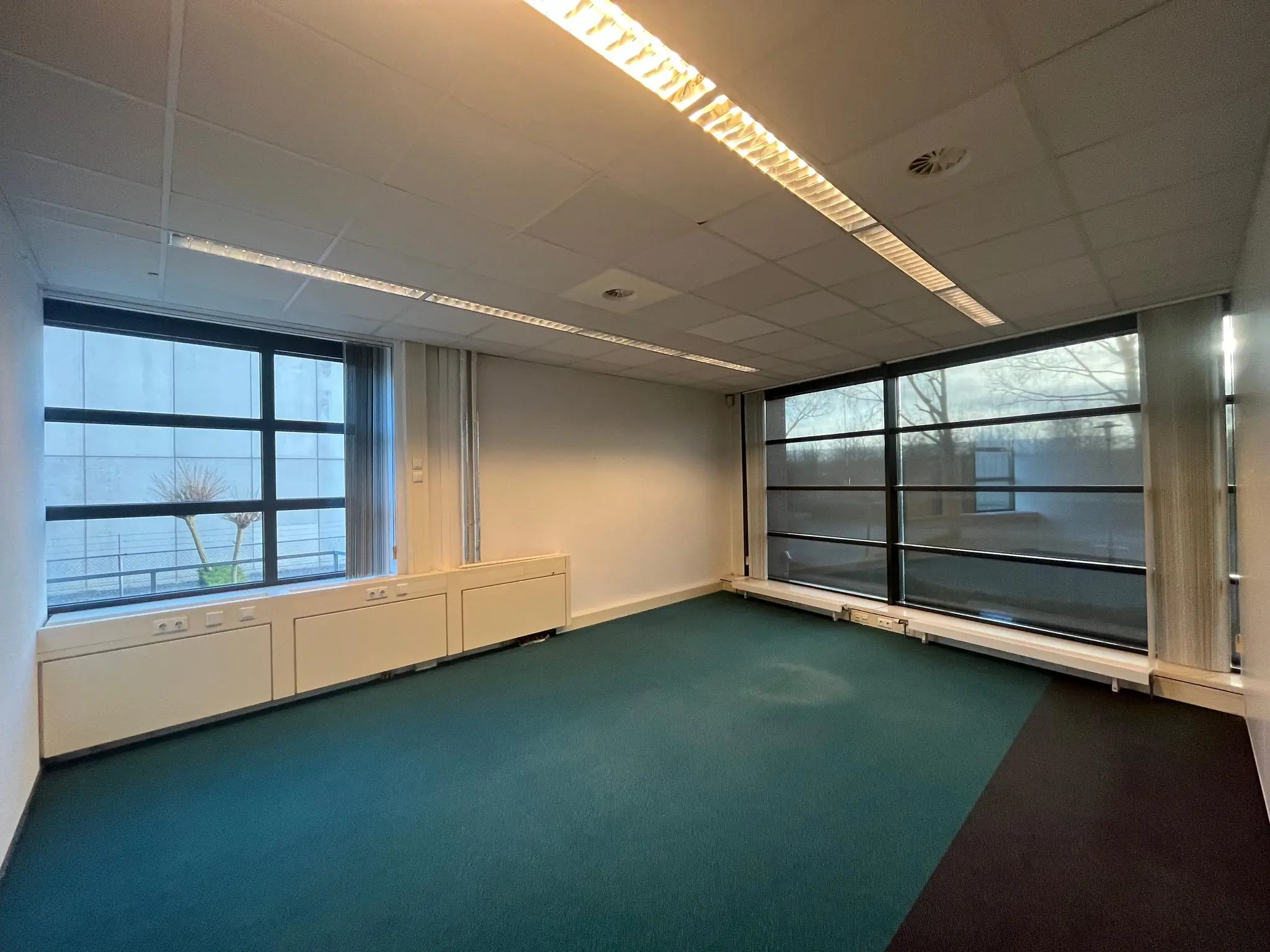 Lege kantoorruimte aan de Zonnebaan met grote ramen, systeemplafond en blauw tapijt.