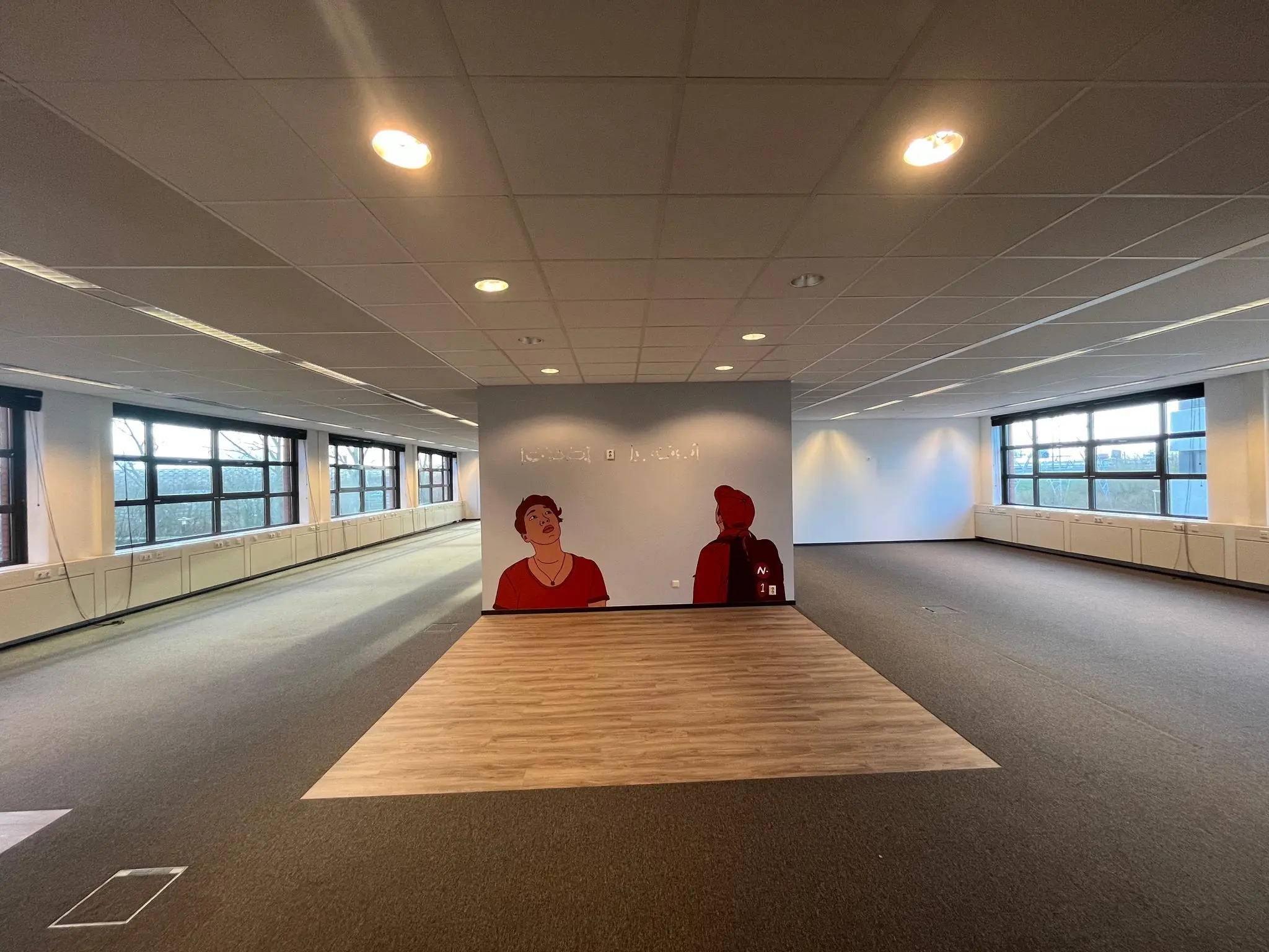 Een lege kantoorruimte met grote ramen en een muurschildering van twee getekende personen op een grijze wand, in het gebouw aan de Zonnebaan.