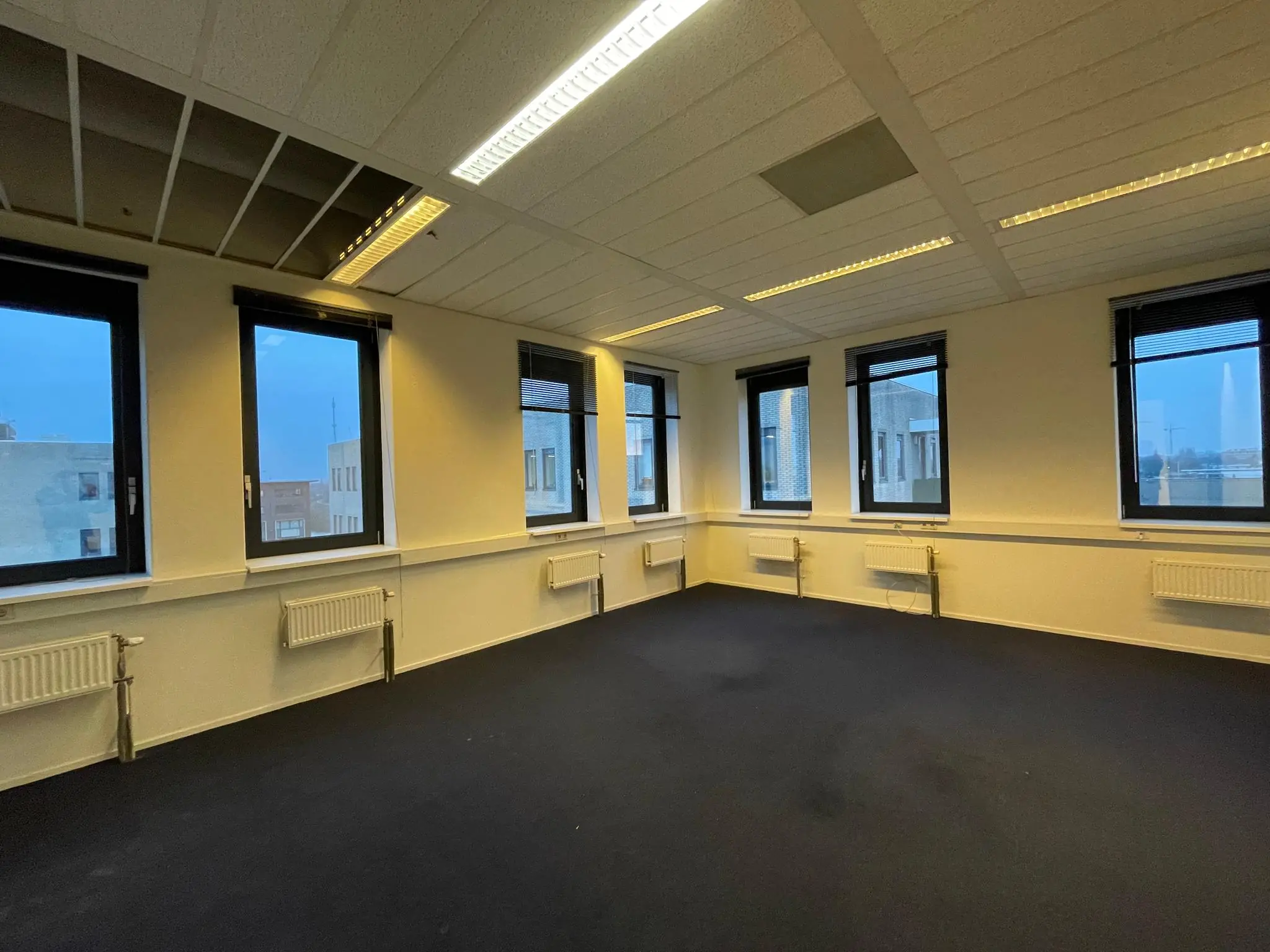 Lege kantoorruimte aan de Zekeringstraat met meerdere ramen, lichtgele muren en donkerblauwe vloerbedekking.