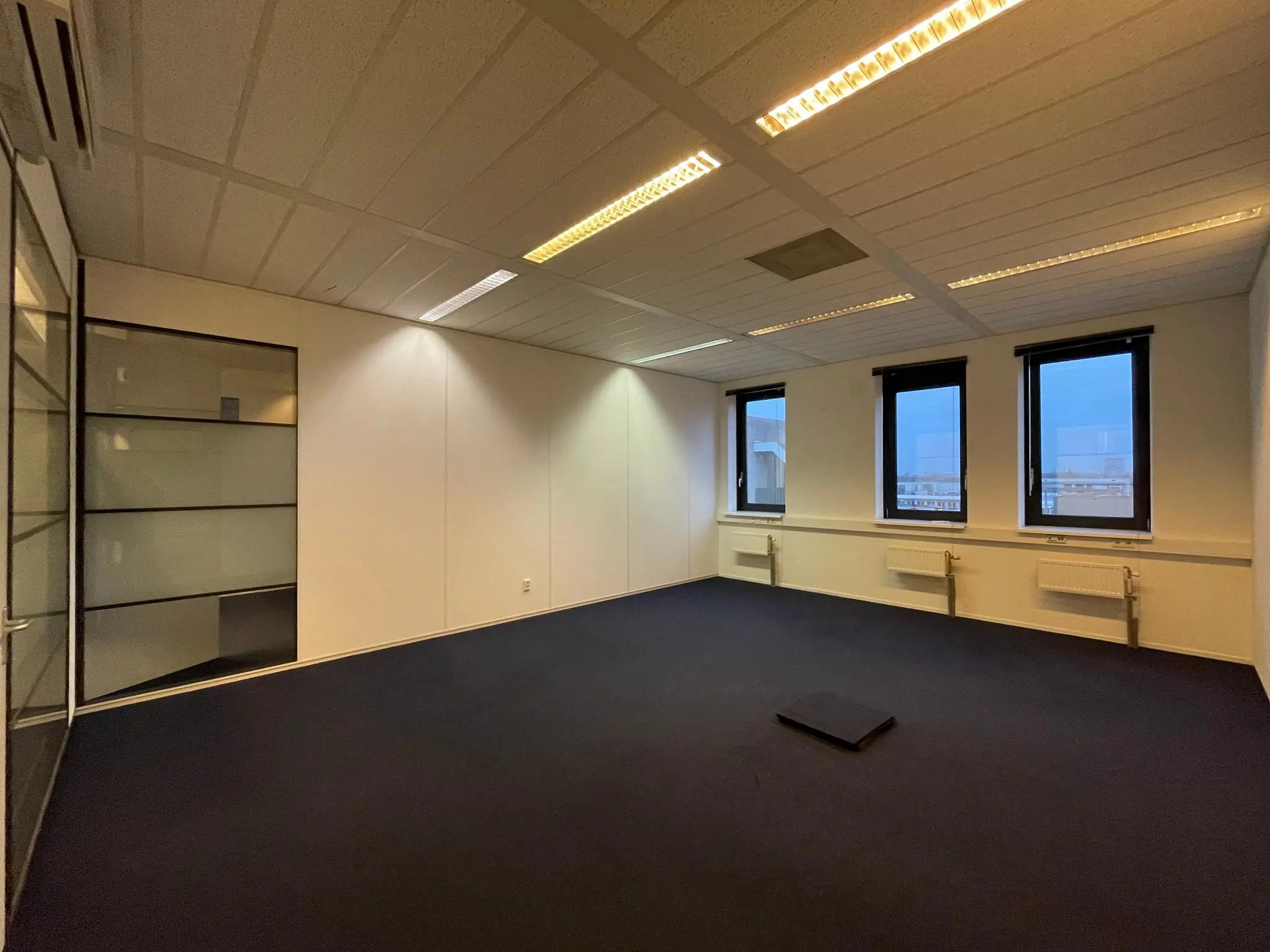 Leeg kantoorruimte in Zekeringstraat met drie ramen, glazen scheidingswand en donker tapijt.