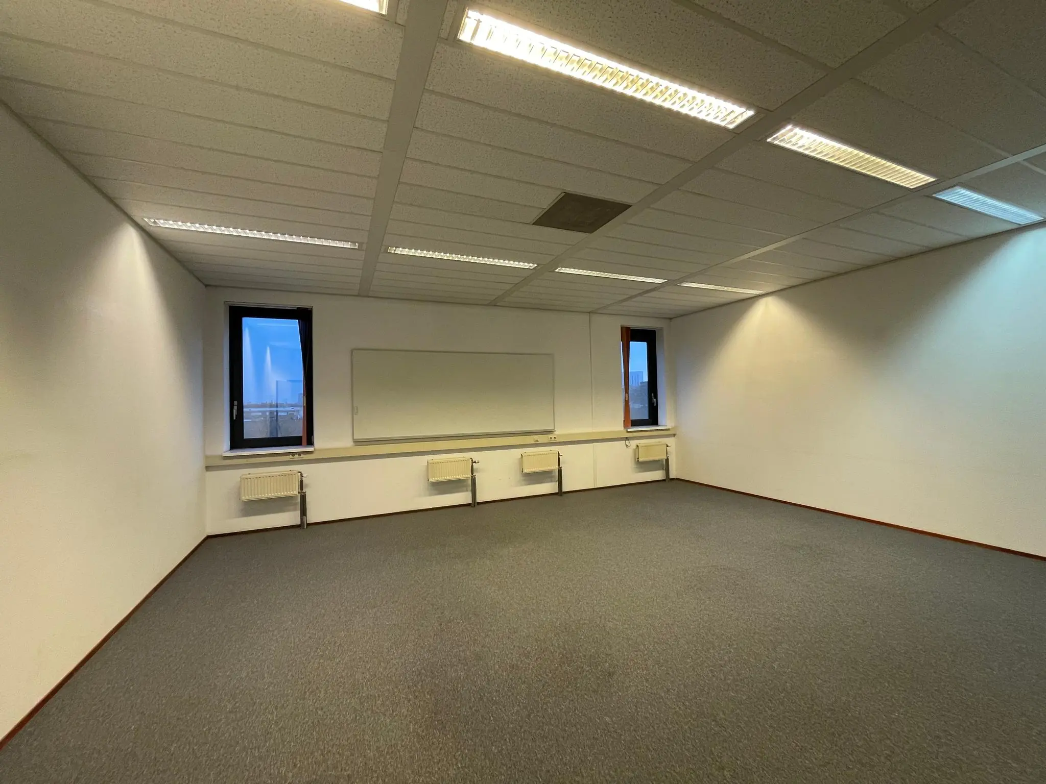 Leeg kantoorruimte aan de Zekeringstraat met wit bord, tapijtvloer en twee ramen.