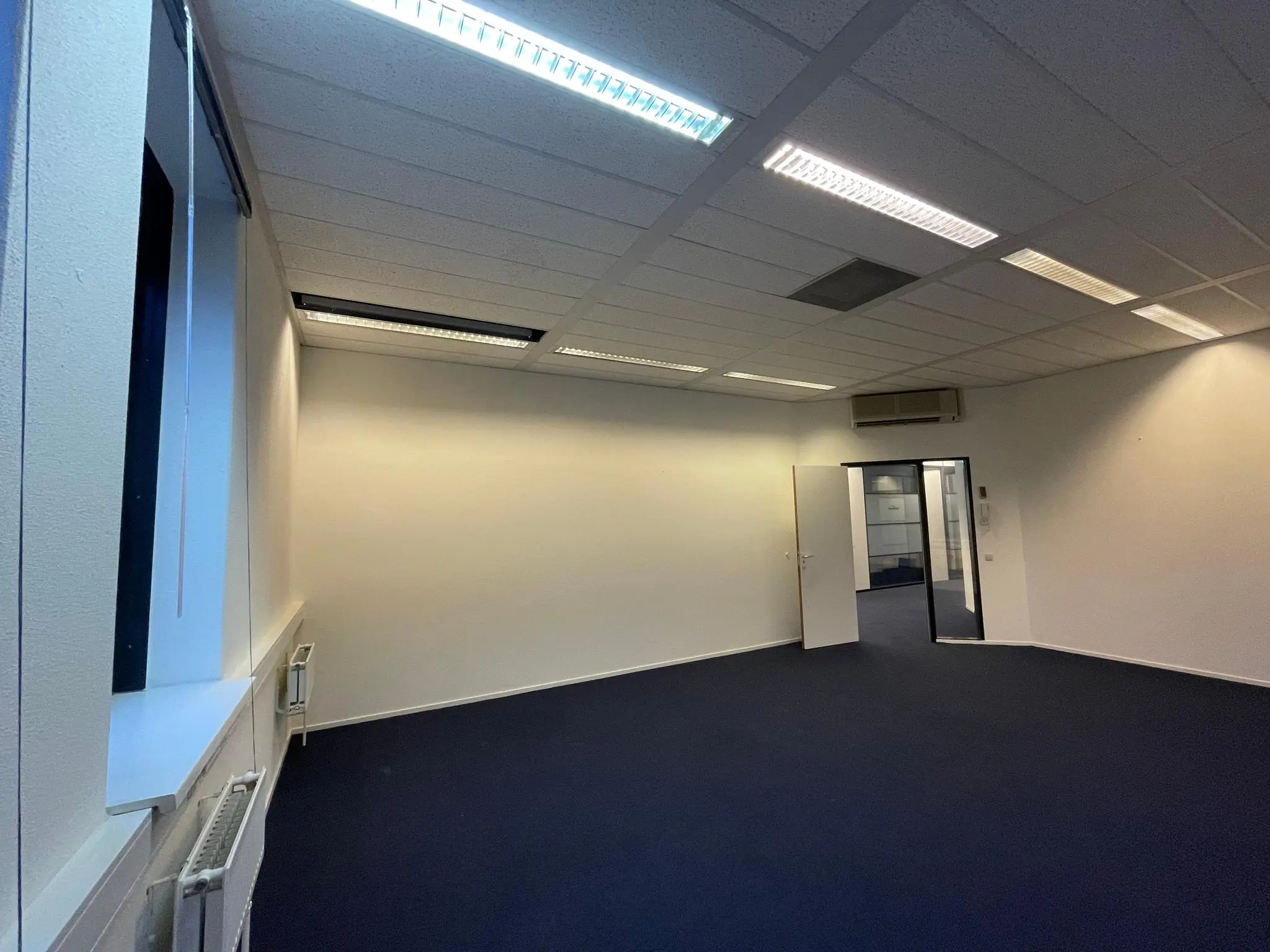 Leeg kantoorruimte aan de Zekeringstraat met blauw tapijt, witte muren en systeemplafond met TL-verlichting.