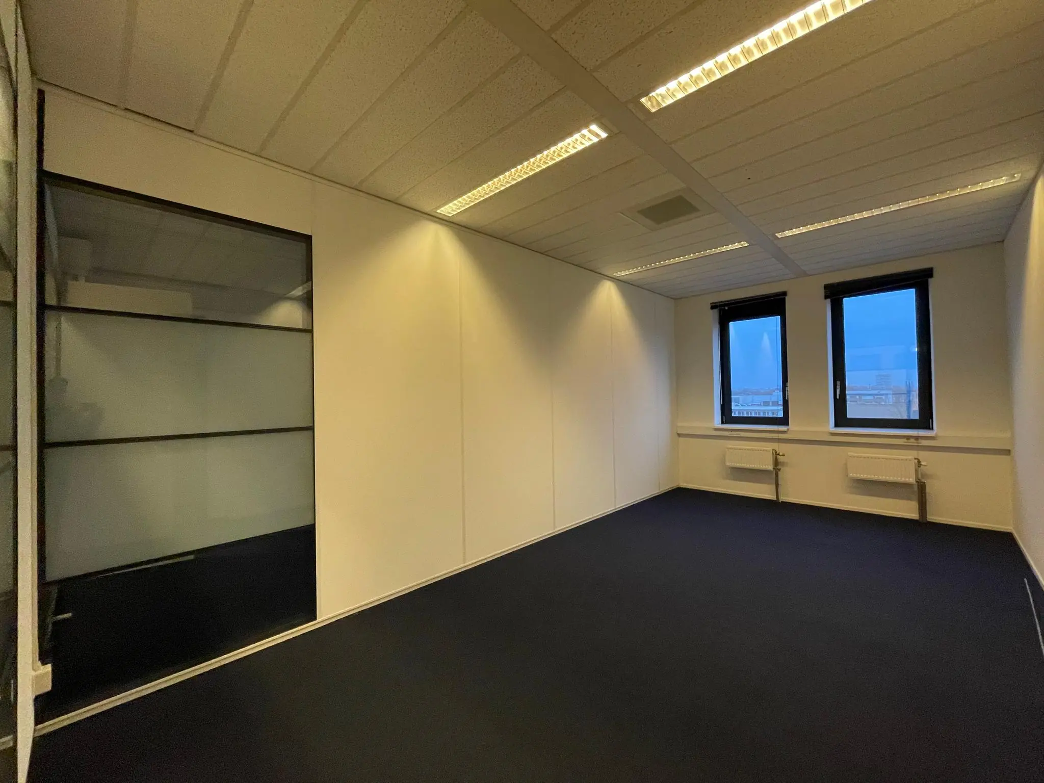 Leeg kantoor met donker tapijt, wit plafond, twee ramen en een glazen wand aan de Zekeringstraat.