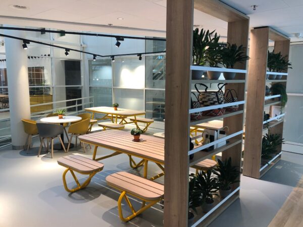 Moderne kantoorlounge aan de Newtonlaan met houten zitbanken, gele accenten en groene planten in open houten stellingen.
