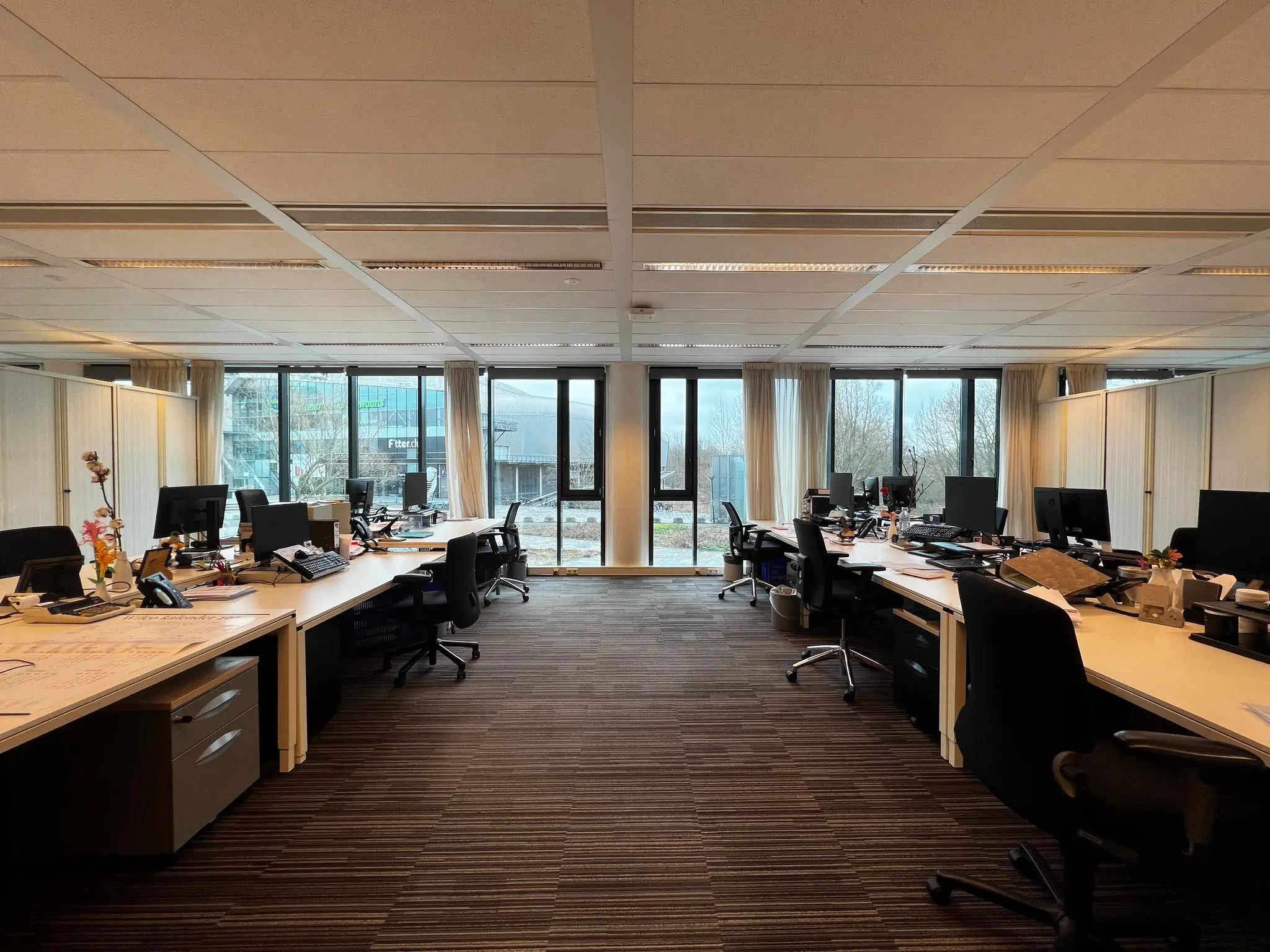 Ruim en modern kantoorpand aan het Herculesplein met werkplekken, bureaus en computers, en uitzicht op buiten.