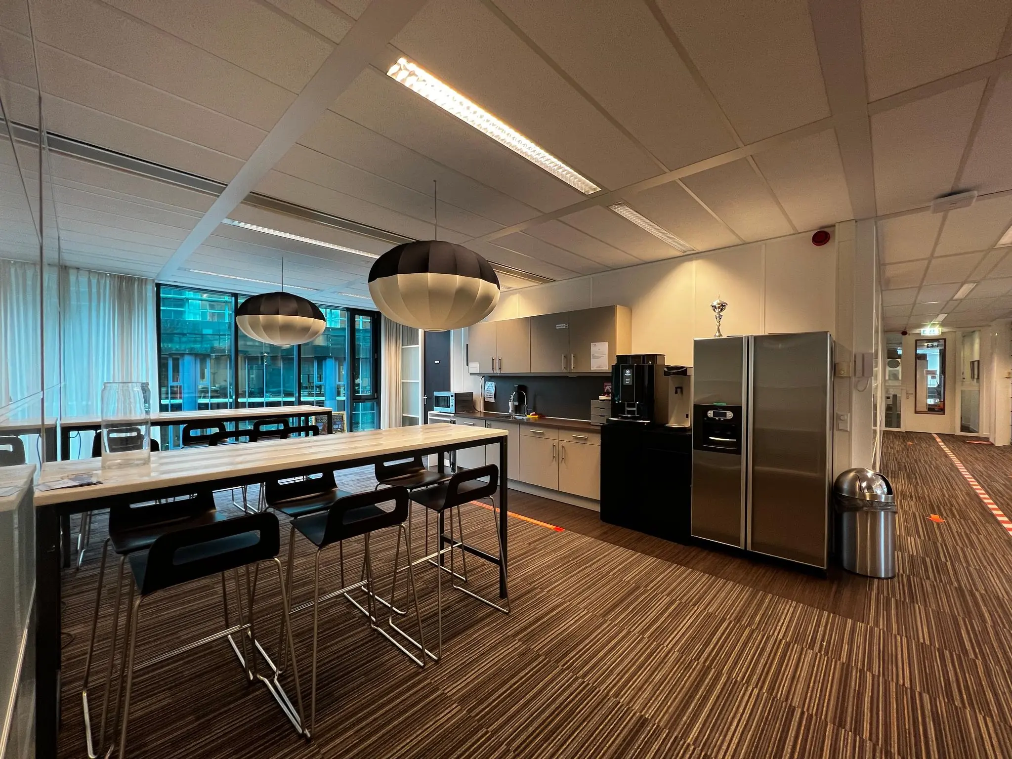 Moderne kantoorkeuken op Herculesplein met een lange eettafel, barkrukken, koffieapparaat en twee koelkasten.