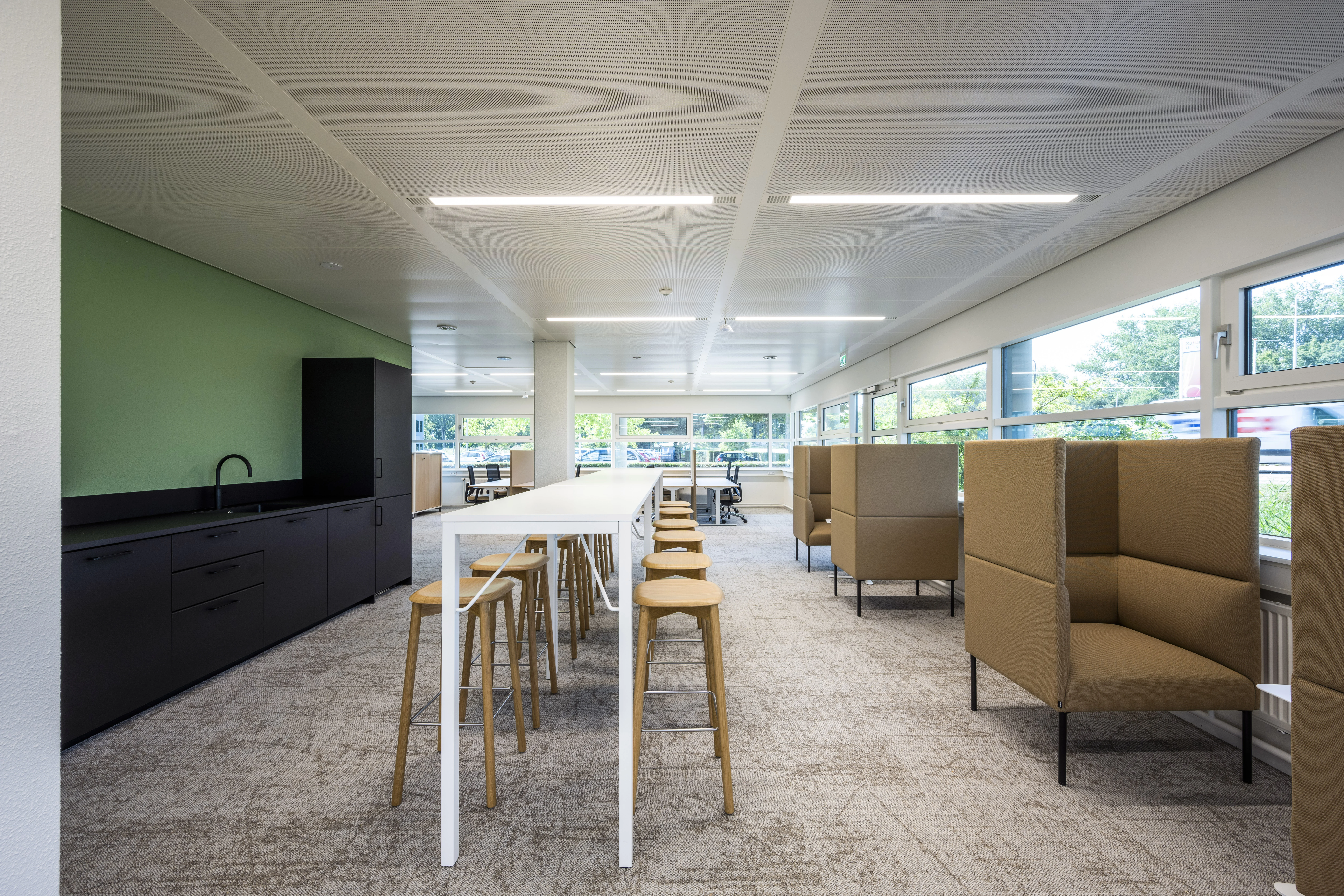 Moderne kantoorruimte met een lange werktafel, barkrukken, een pantry en comfortabele zitnissen langs de ramen.