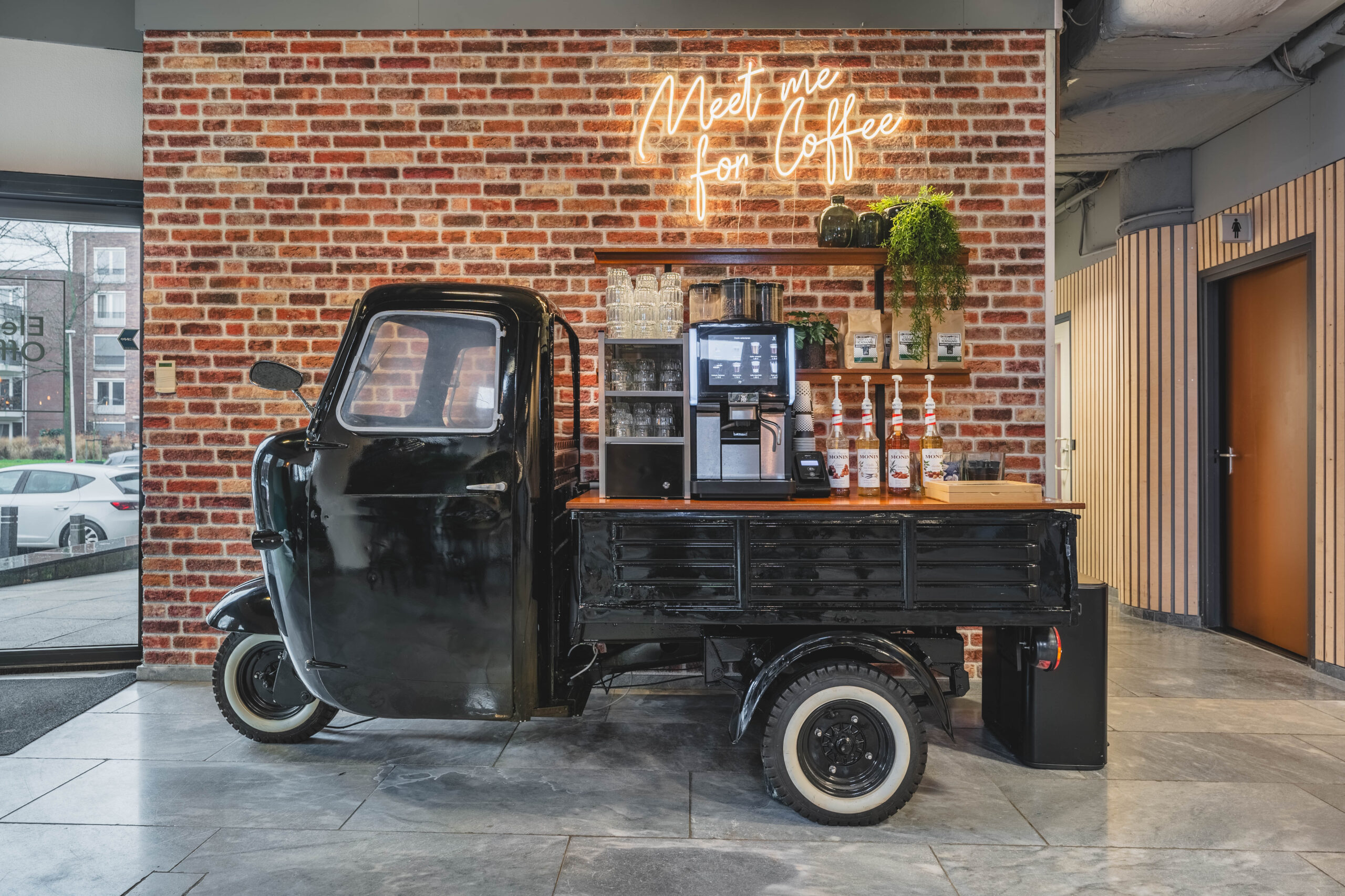 Een mobiele koffiebar in een gemodificeerde driewieler met een bakstenen muur op achtergrond en een neonbord "Meet me for Coffee".