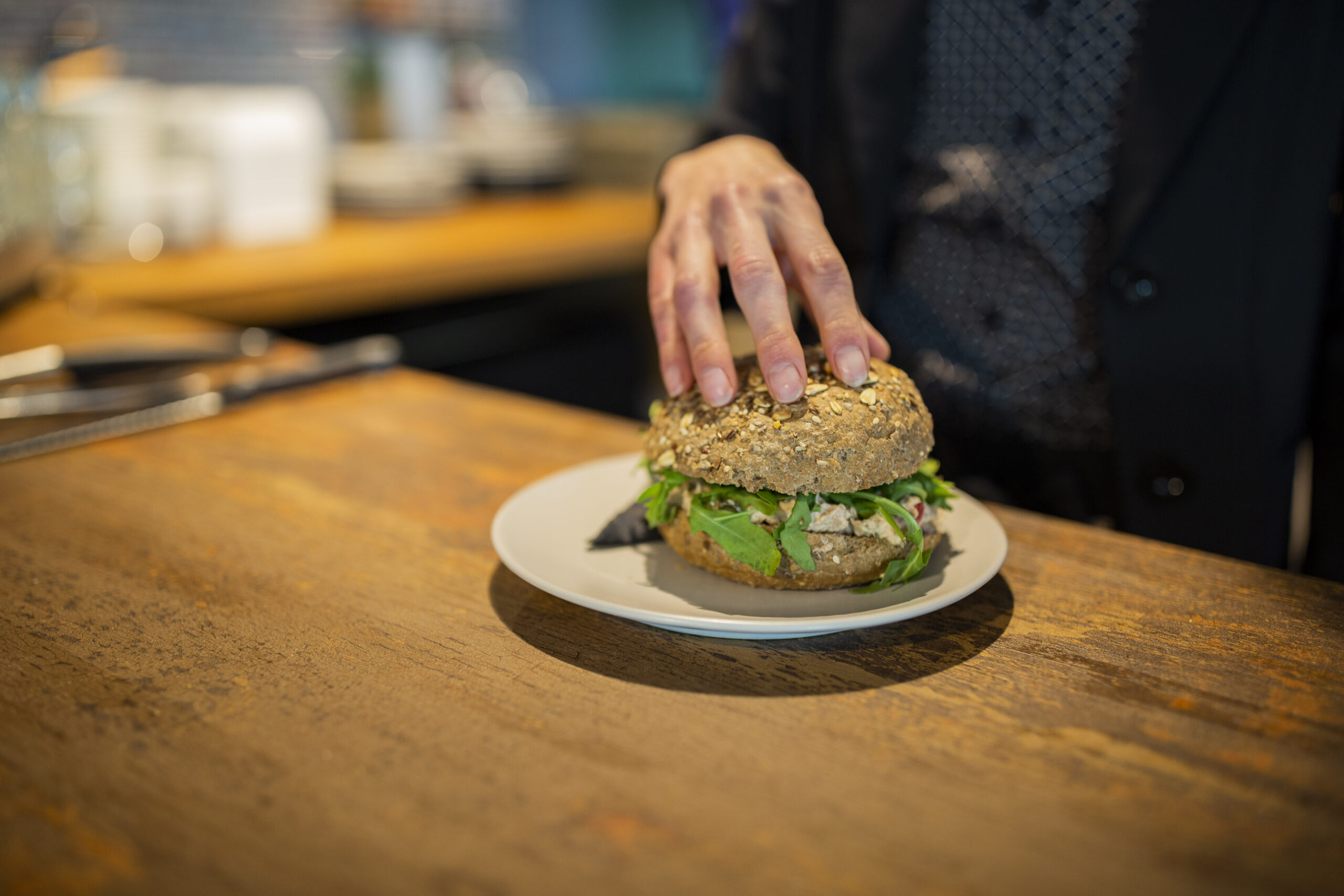 Een persoon pakt een komkommer- en bladgroente-sandwich op een wit bord op een houten tafel.