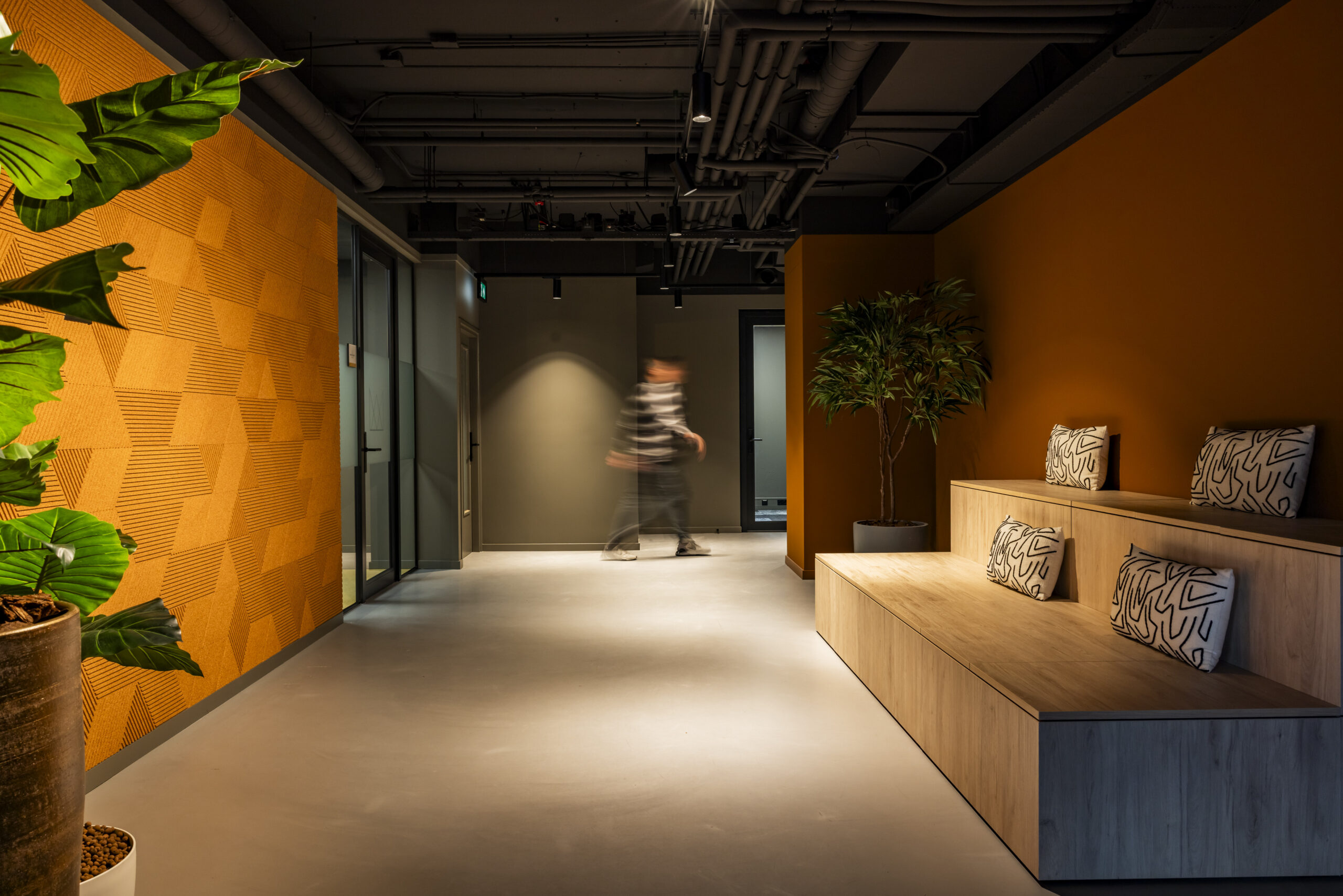 Een moderne gang in een kantoor met een houten zitgedeelte, sierkussens, planten en een geometrische oranje muur.