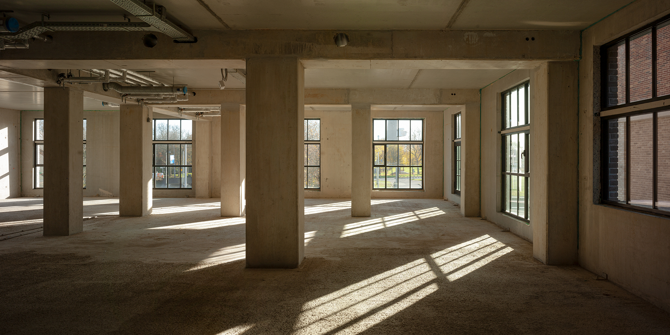Een leeg, zonovergoten interieur met betonpilaren en grote ramen langs de Karl Weisbardstraat.